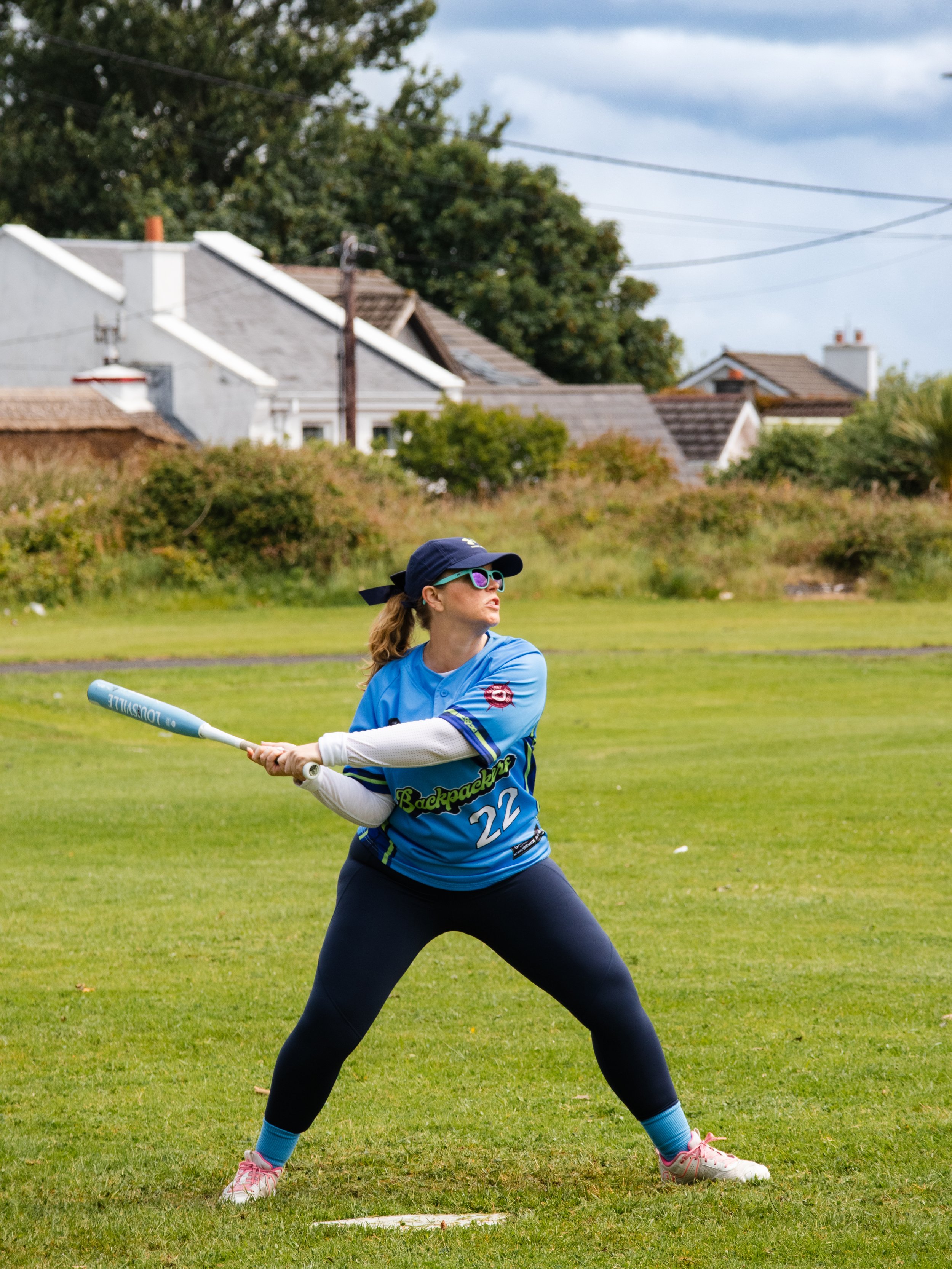 A woman playing softball outdoors, wearing a blue sports jersey, dark leggings, a cap, and sunglasses, holding a bat ready to swing on a grassy field with houses and trees in the background.