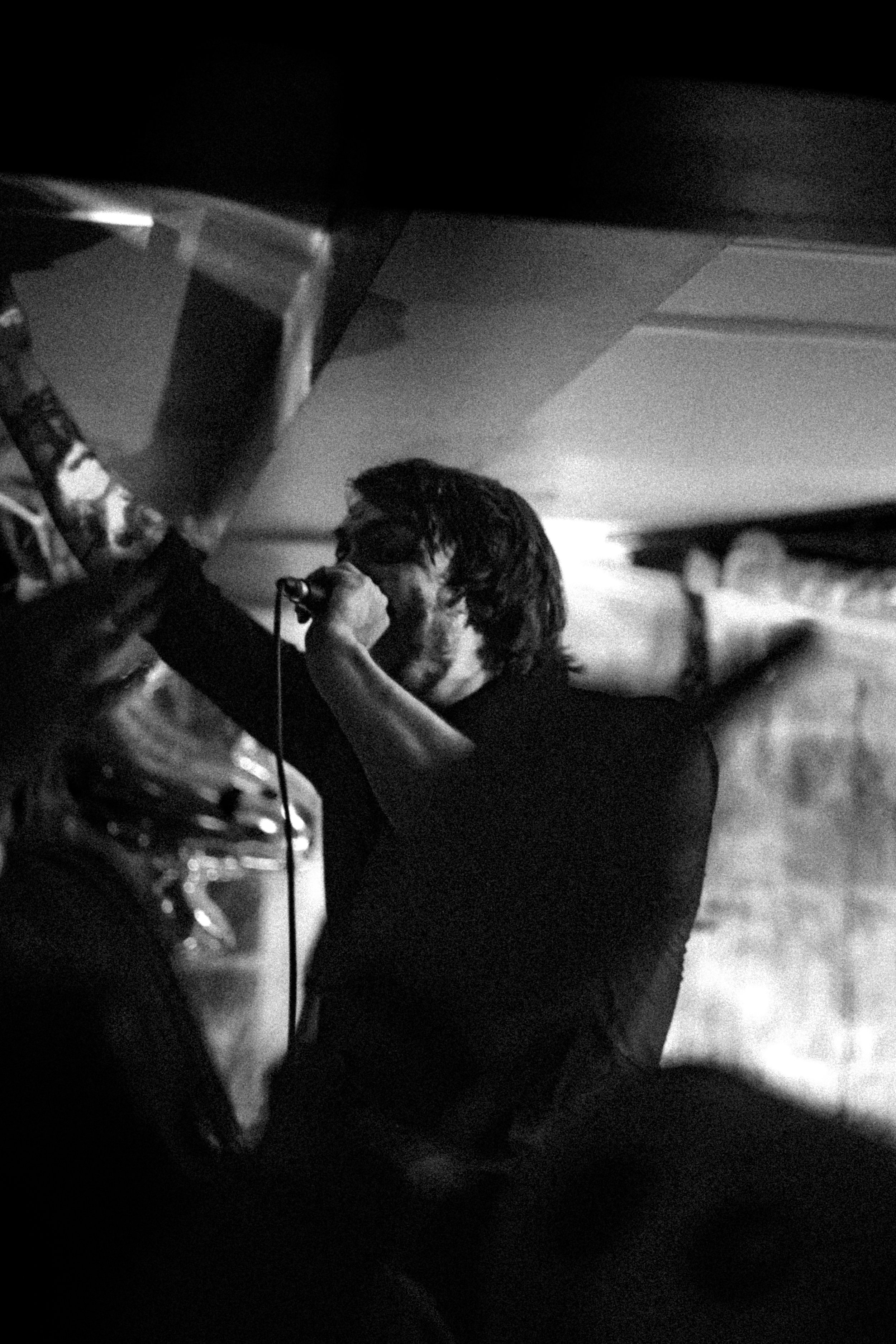A black and white photo of a man singing into a microphone on stage with his arm raised, in a dark setting.