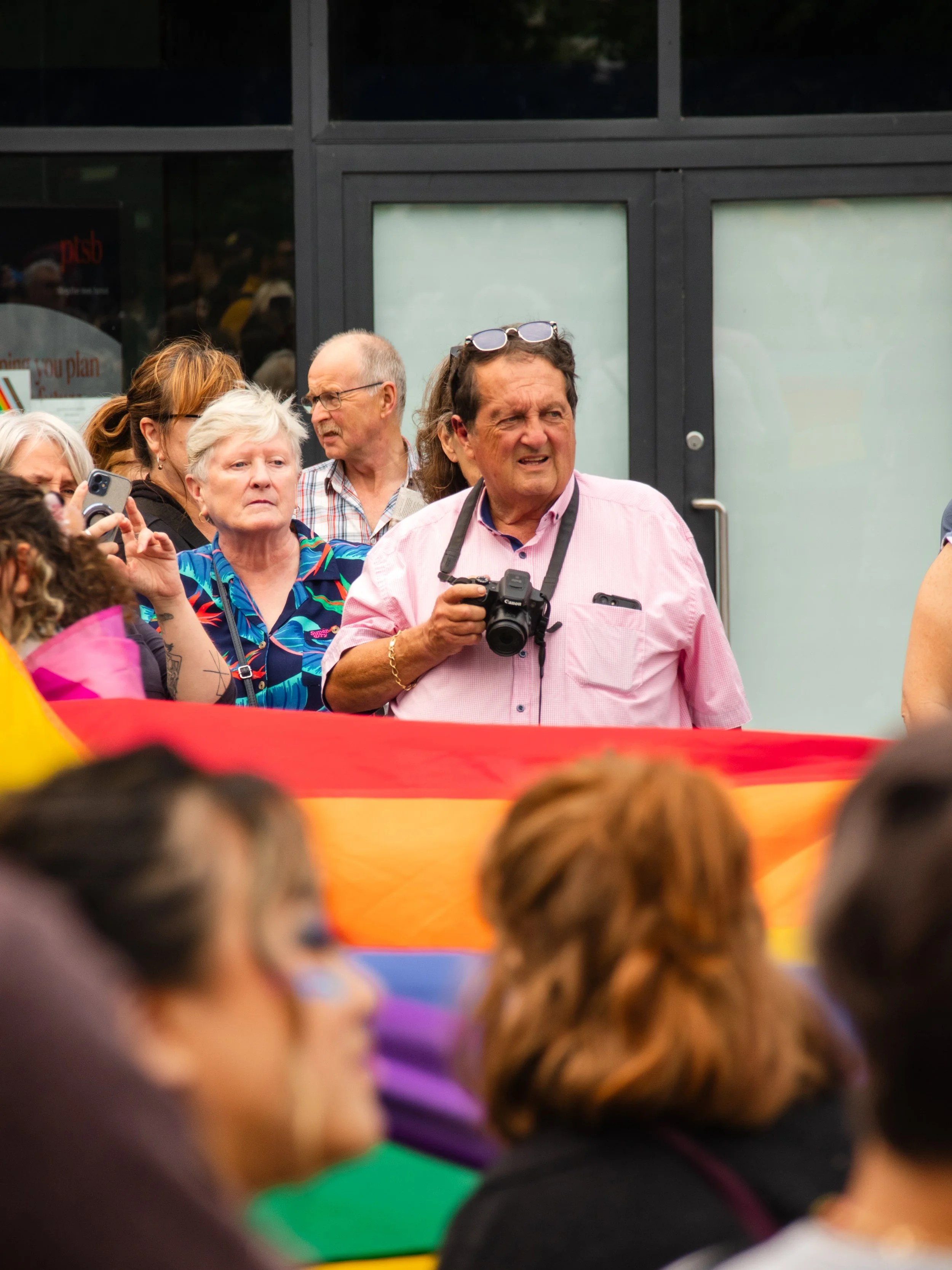A group of diverse people at a rally or event, holding a rainbow LGBTQ+ pride flag. The crowd is densely gathered and appears engaged, with some people taking photos and others watching. The background shows a building with black framing and frosted 