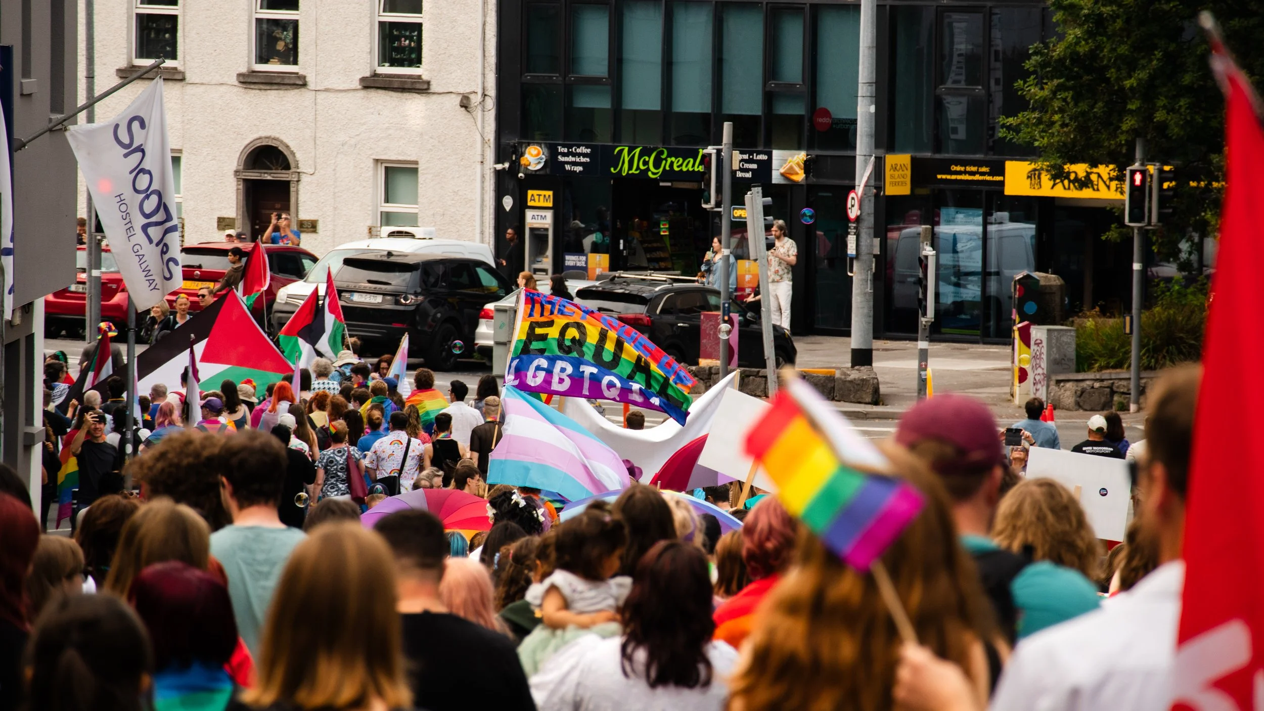 Crowd of people participating in a pride march with rainbow flags and banners, standing on a city street with buildings and shops in the background.