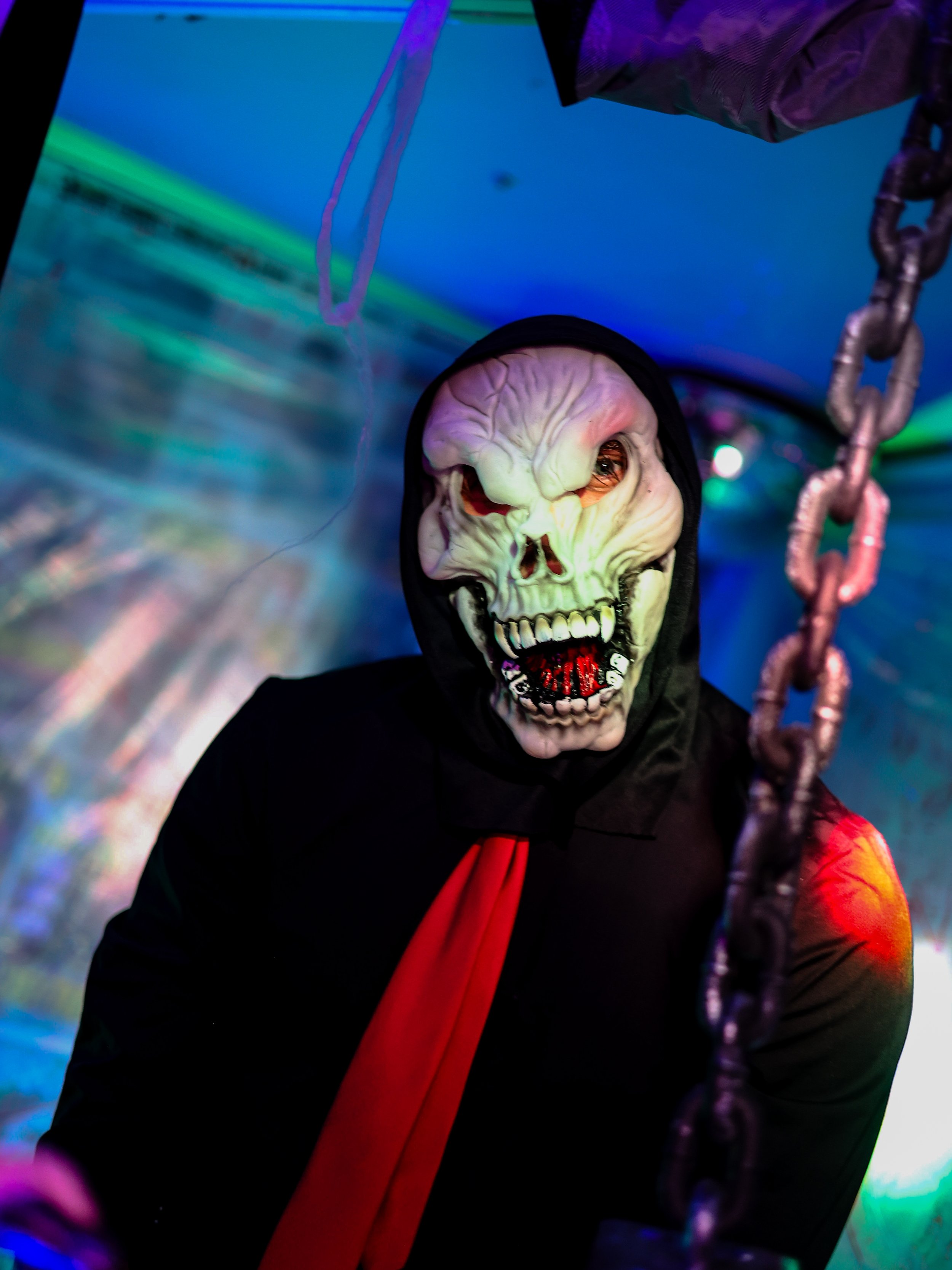 Person in a spooky skeleton mask wearing a black hooded cloak and red cape, surrounded by colorful neon lights.