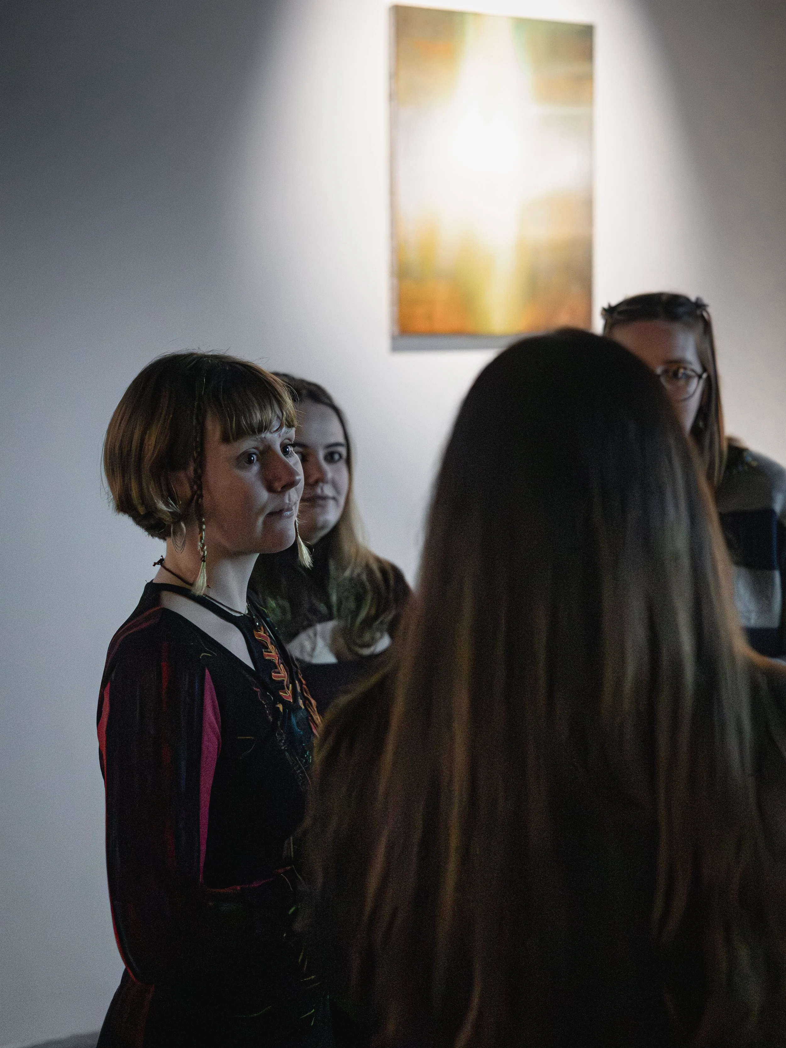 Group of women standing indoors, with one woman in the foreground wearing a black dress with colorful patterns, and an abstract painting hanging on the wall behind them.