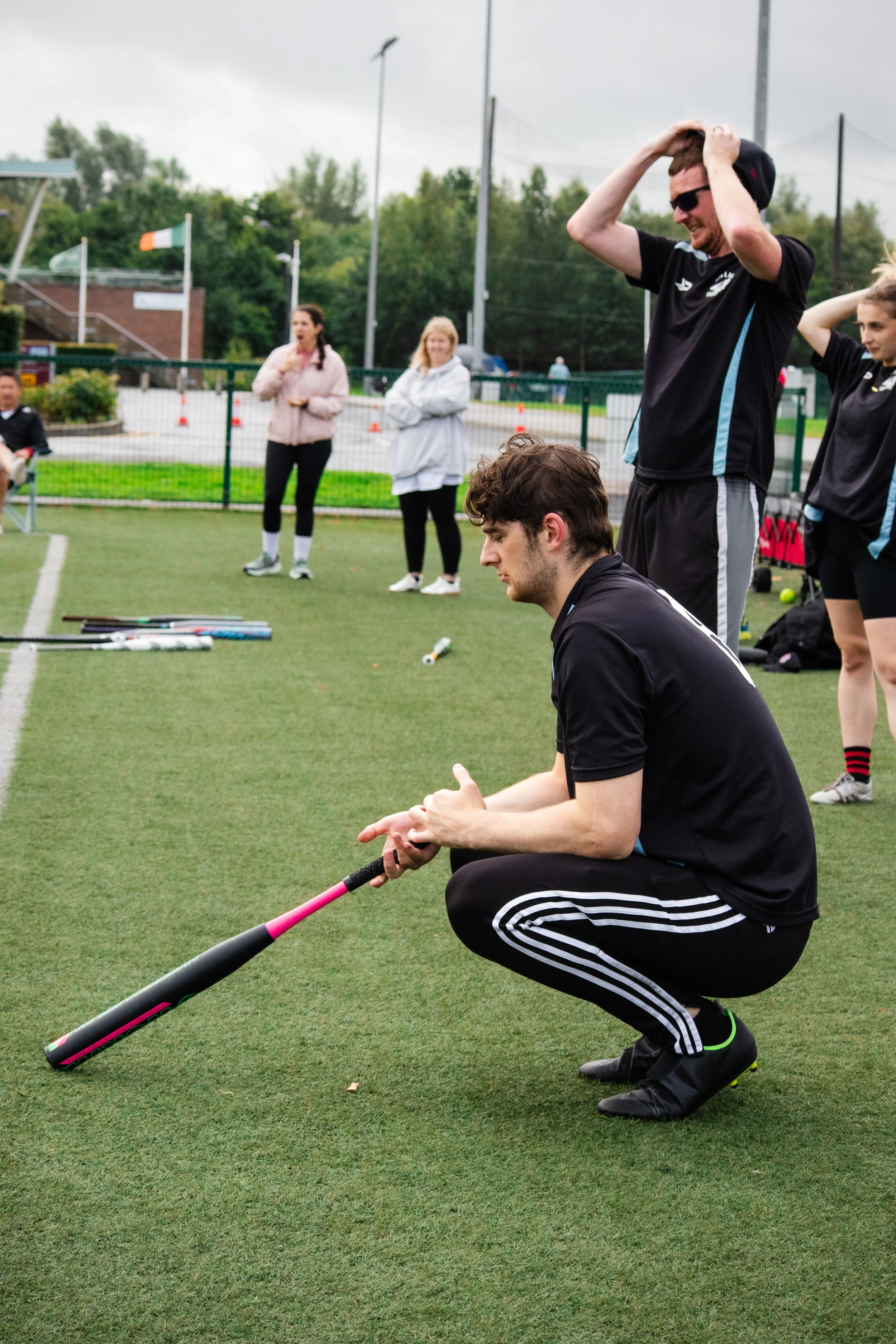 A group of people on a sports field, with one young man squatting and holding a cricket bat, surrounded by others standing and watching during daytime.