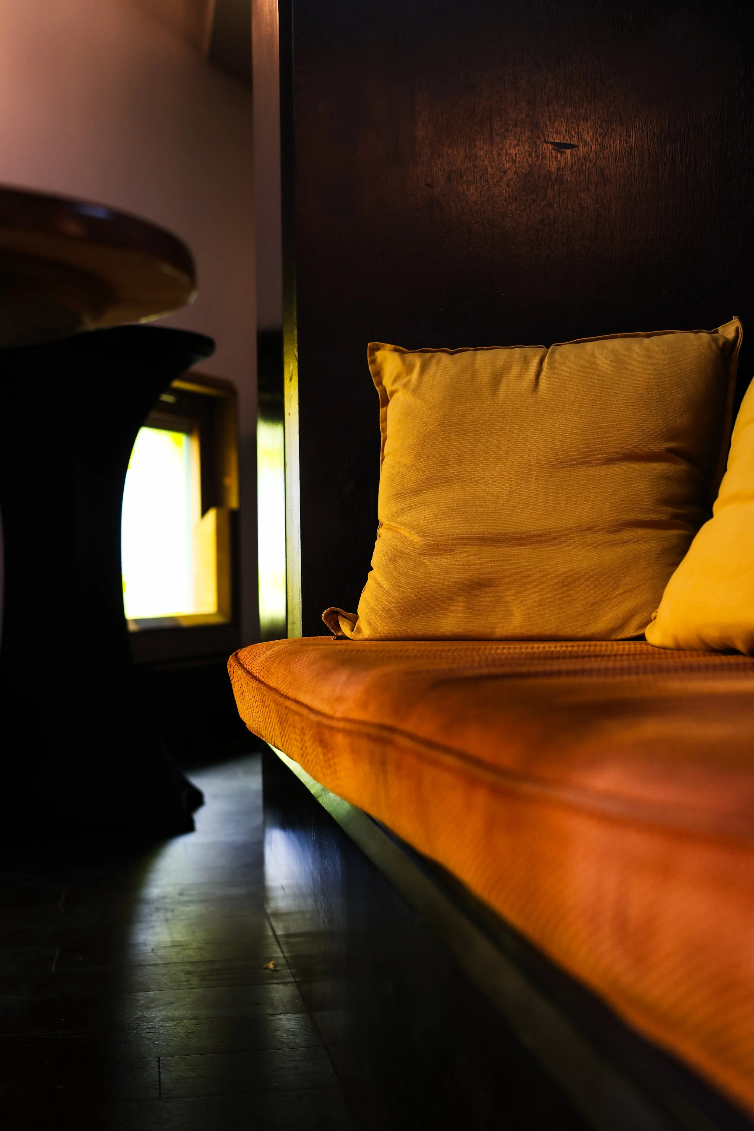 Close-up of an orange cushioned bench with a beige pillow, in a dimly lit room with a small window in the background.
