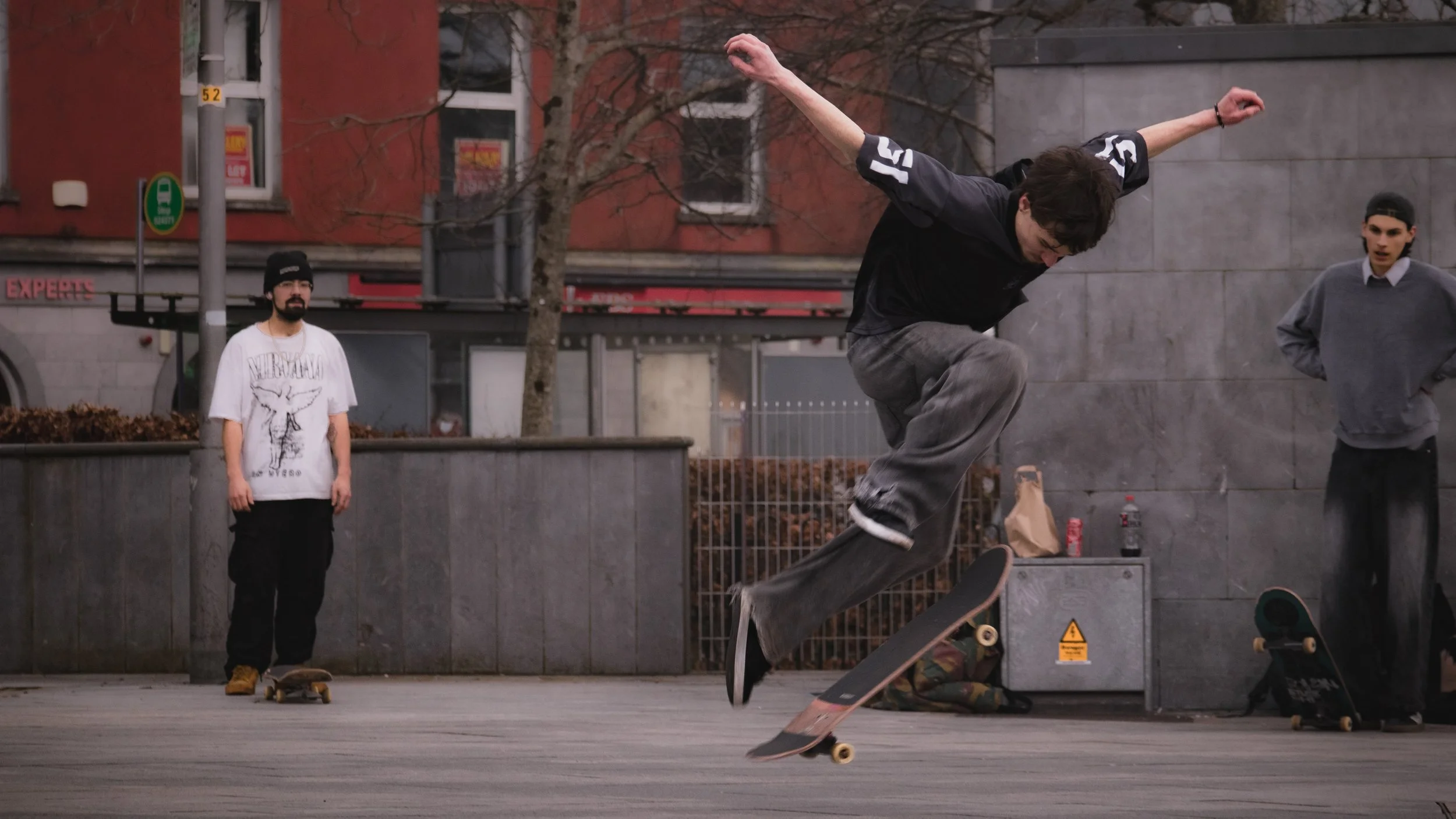 A young man skateboarding in an outdoor urban setting with two other young men watching.