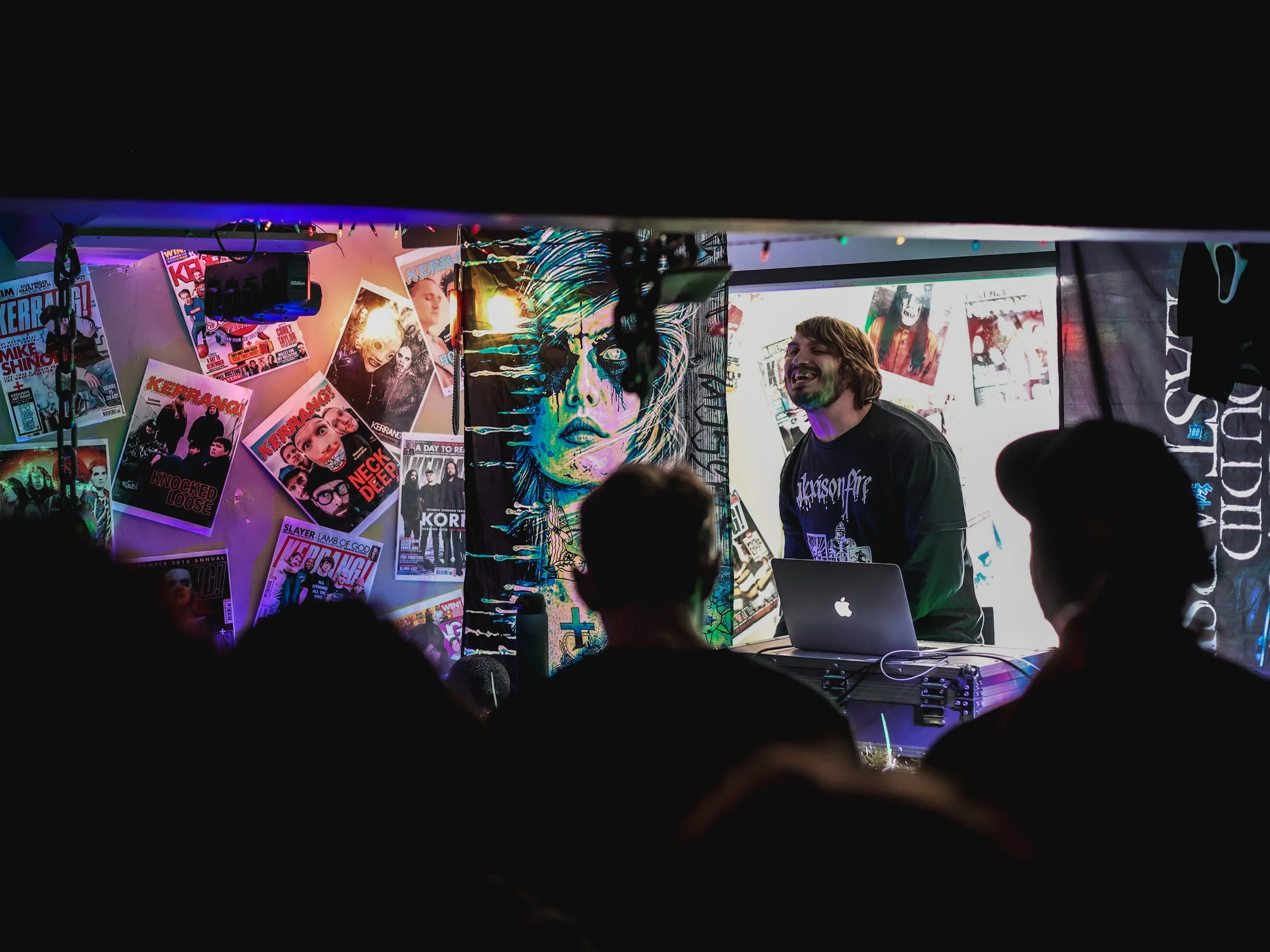 A musician with a beard and long hair is performing at a small indoor venue, standing behind a laptop, surrounded by posters, artwork, and musical equipment, with an audience in the foreground.
