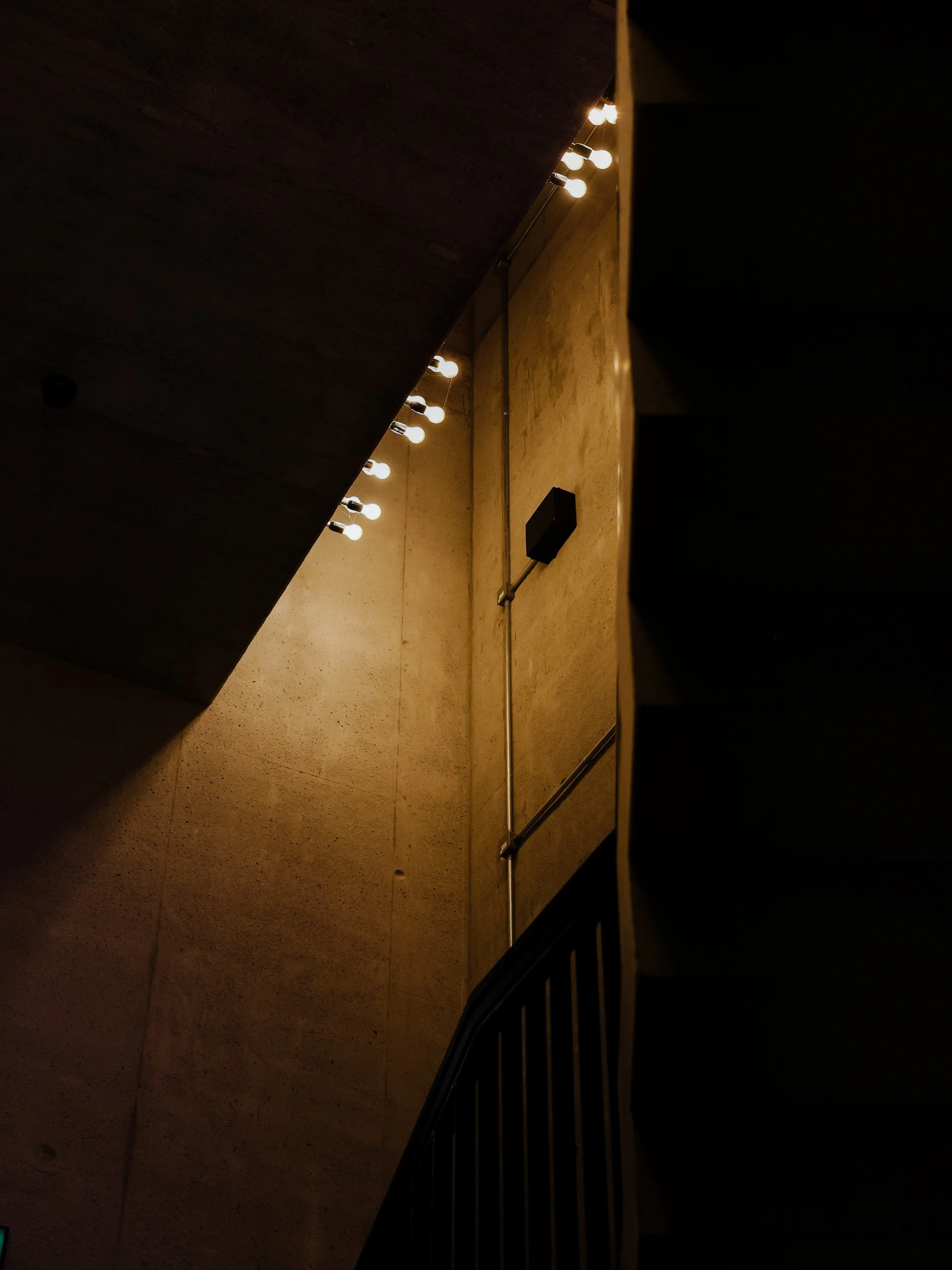 Indoor staircase with a staircase railing in the foreground, a burlap textured wall in the background, and string lights hanging from the ceiling illuminating the space.