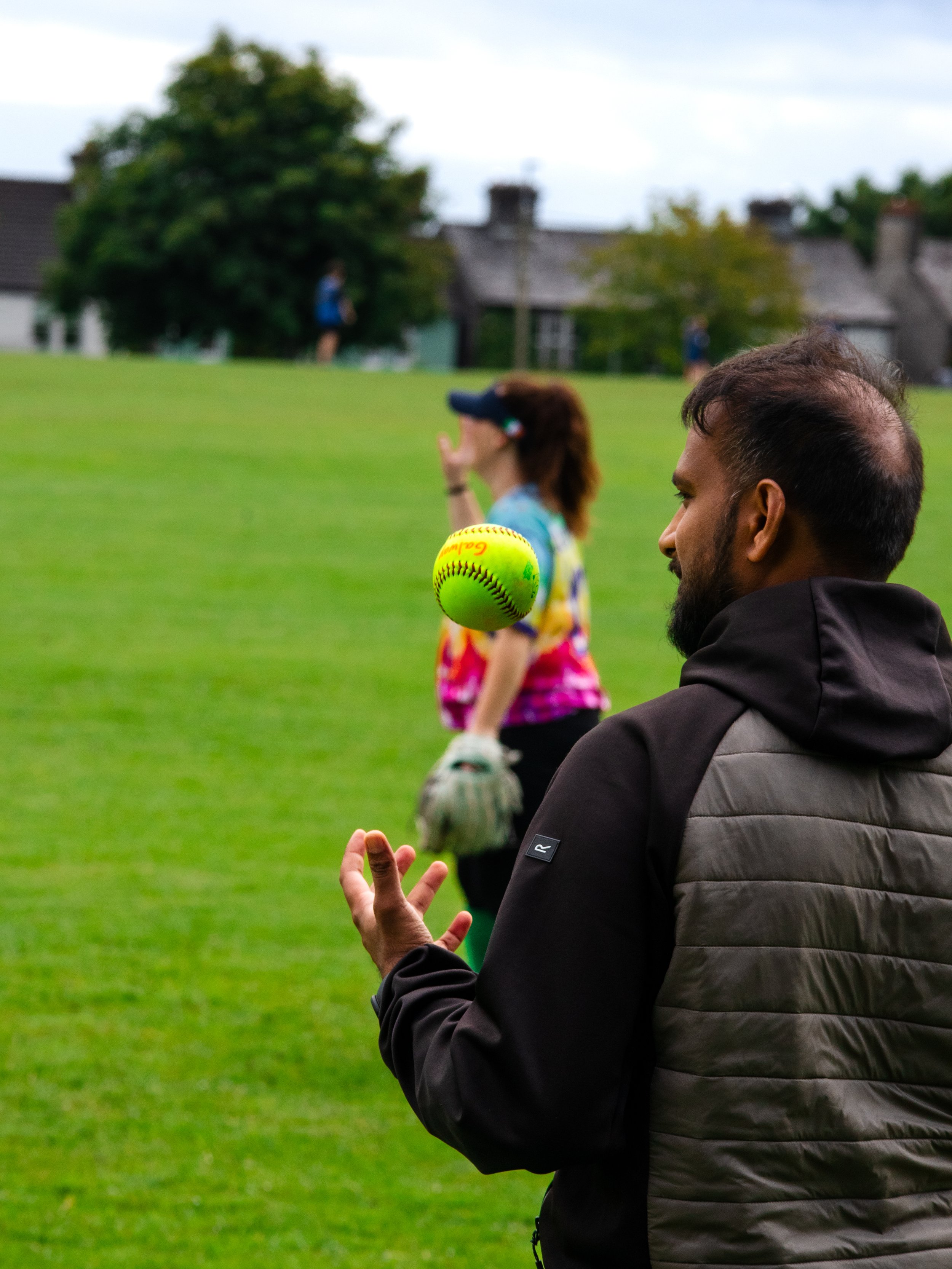 A man in a black jacket tossing a bright yellow softball in the air on a grassy field, with a woman in colorful sports clothing and a cap in the background, holding a softball glove, outdoors in a park-like setting.