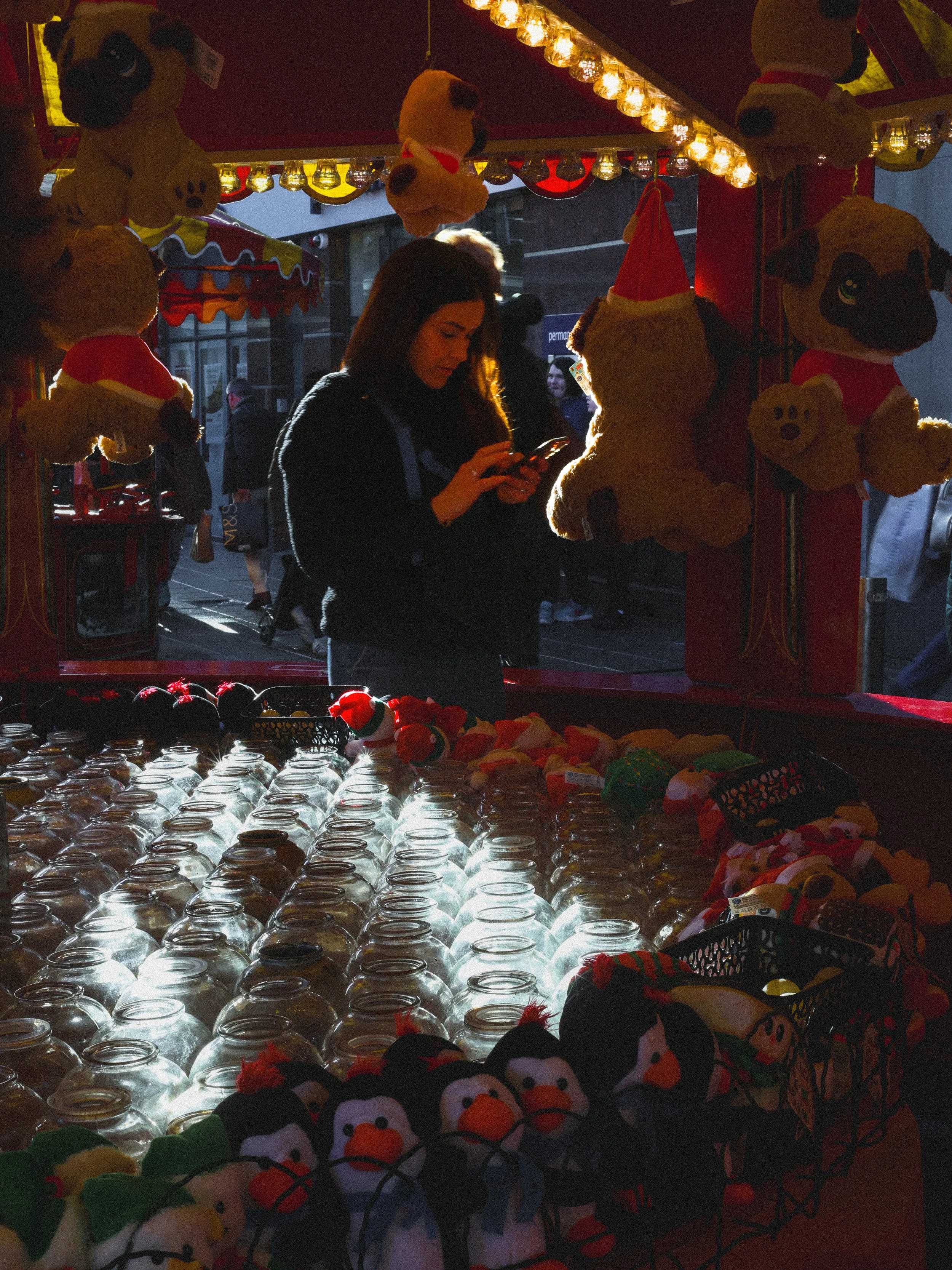 A woman shopping at a holiday-themed fair stall with plush toys and glass jars, illuminated by warm string lights.