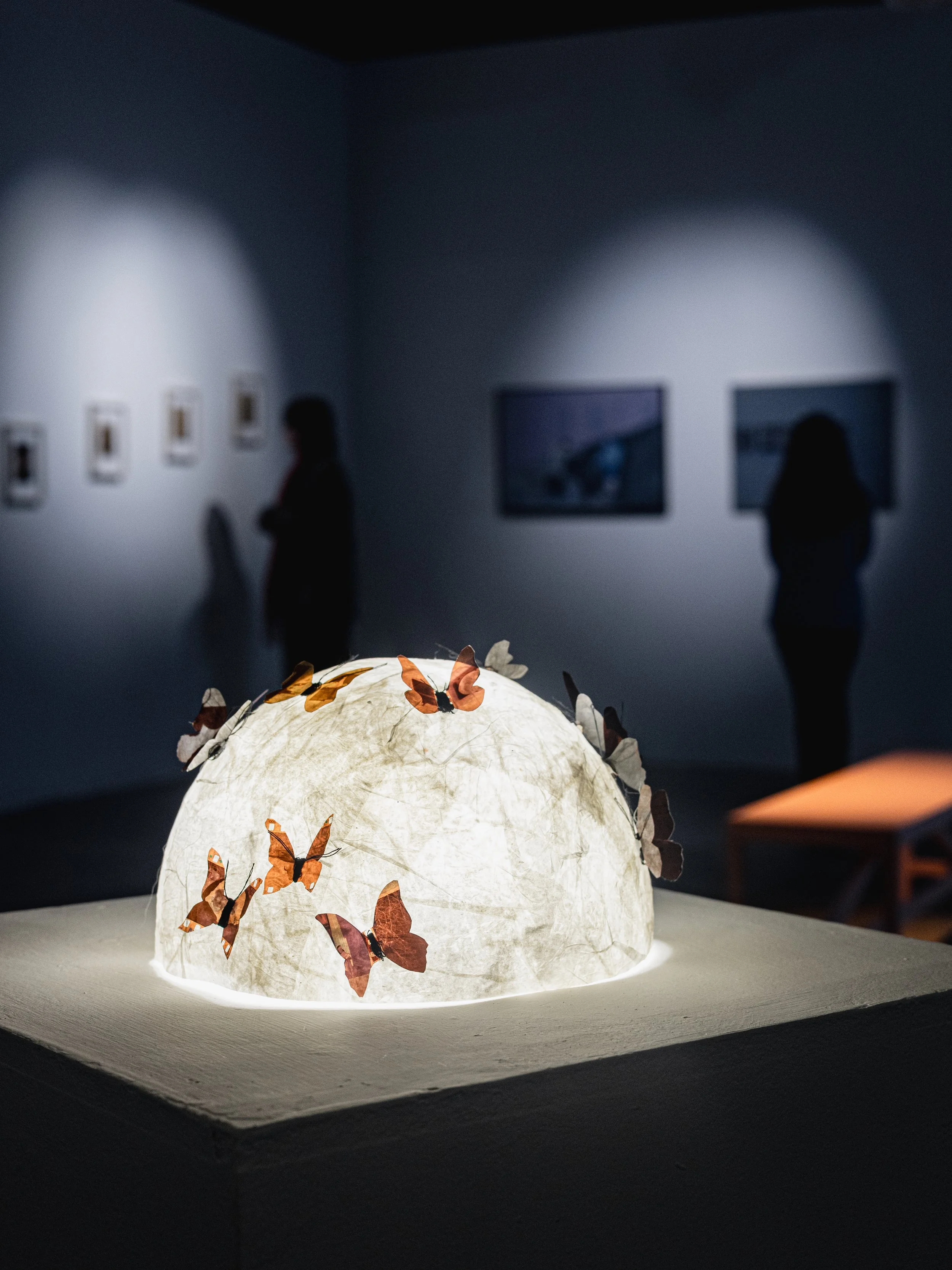 A glowing sculptural piece with butterfly decorations on it, displayed on a pedestal in an art gallery with visitors observing in the background.