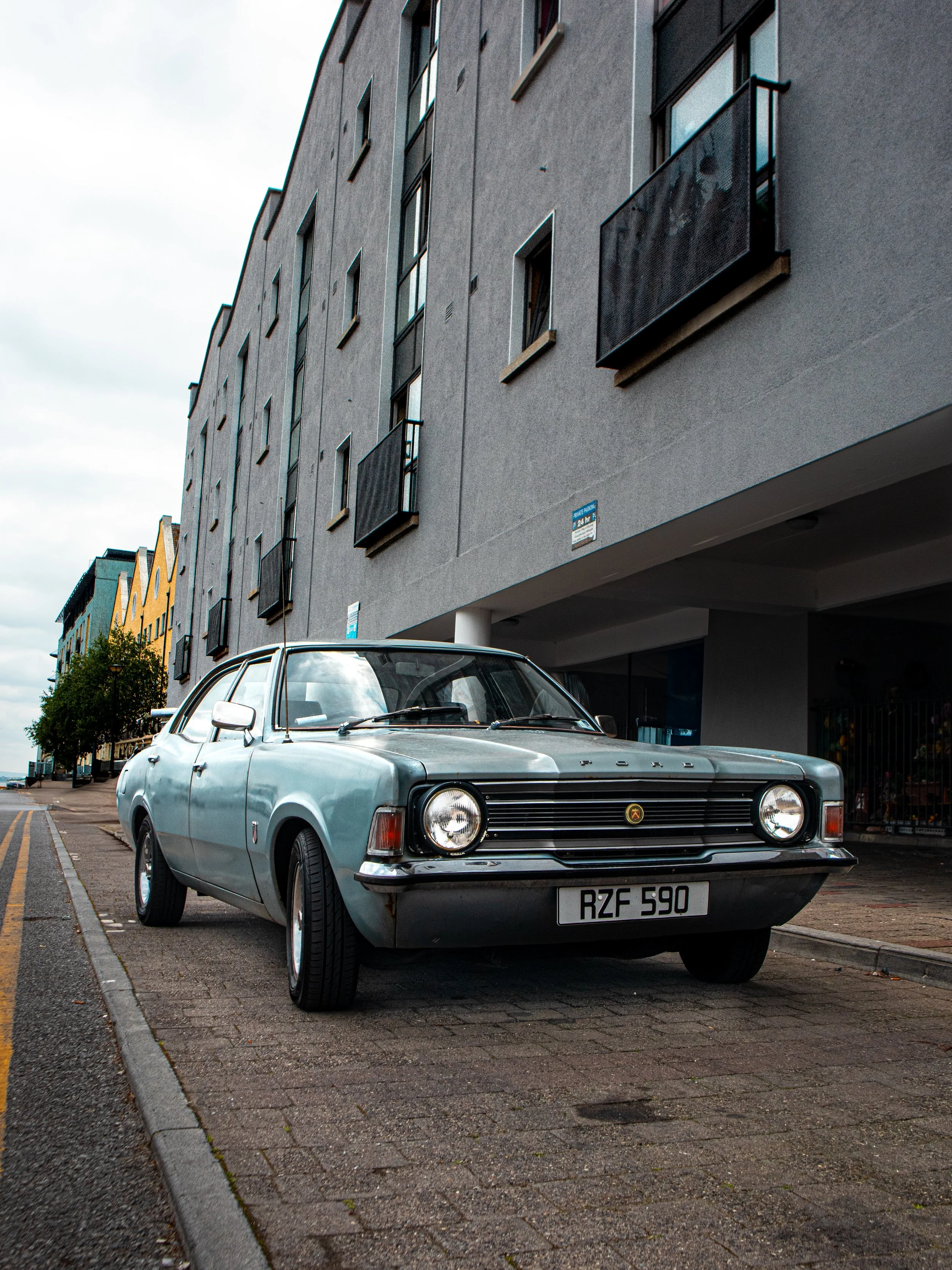 A vintage silver car with the license plate RZF 590 parked on a city street near modern buildings with balconies.