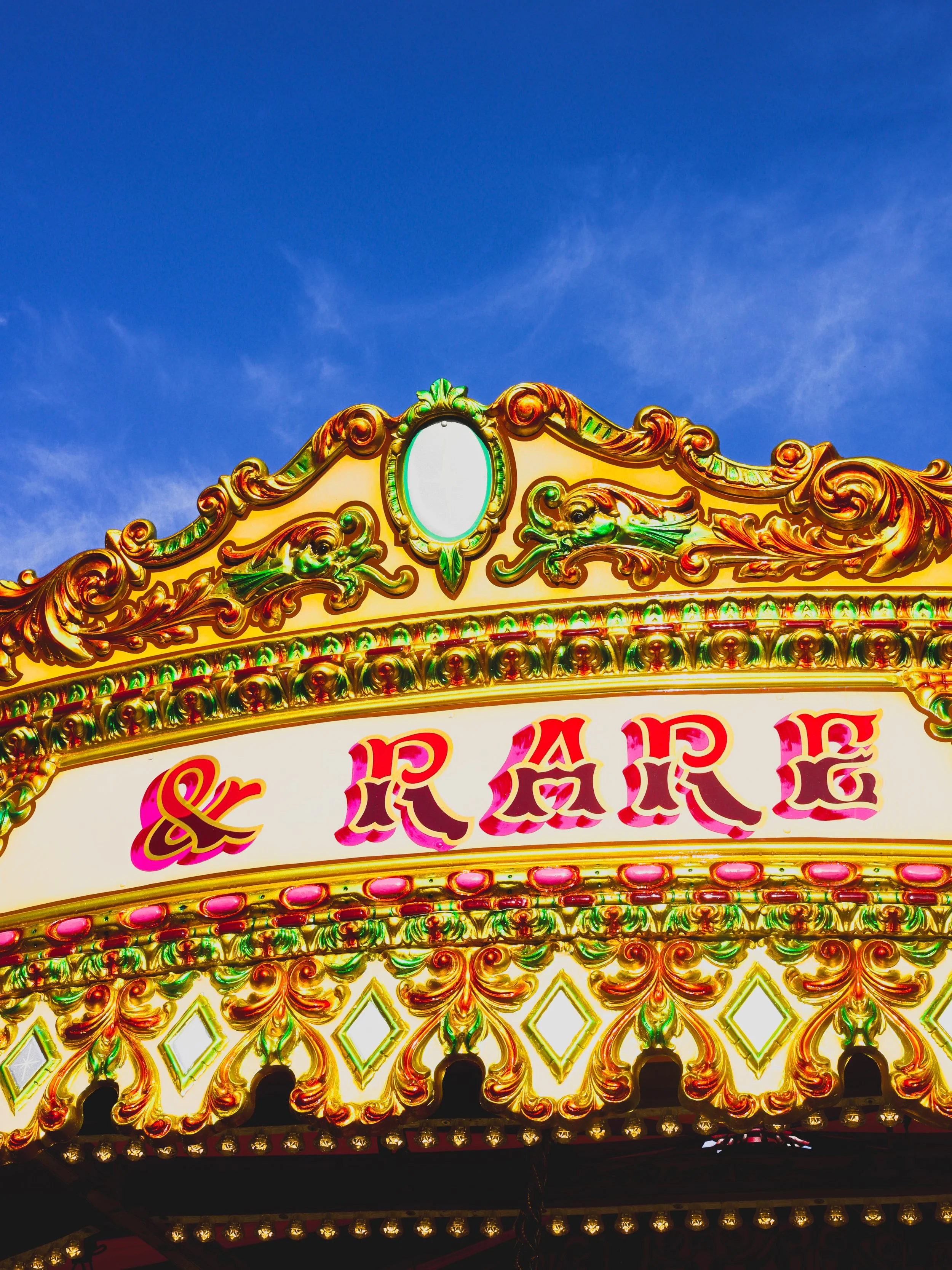 Colorful carnival ride sign with ornate gold trim, pink lettering spelling 'Carousel,' against a blue sky.