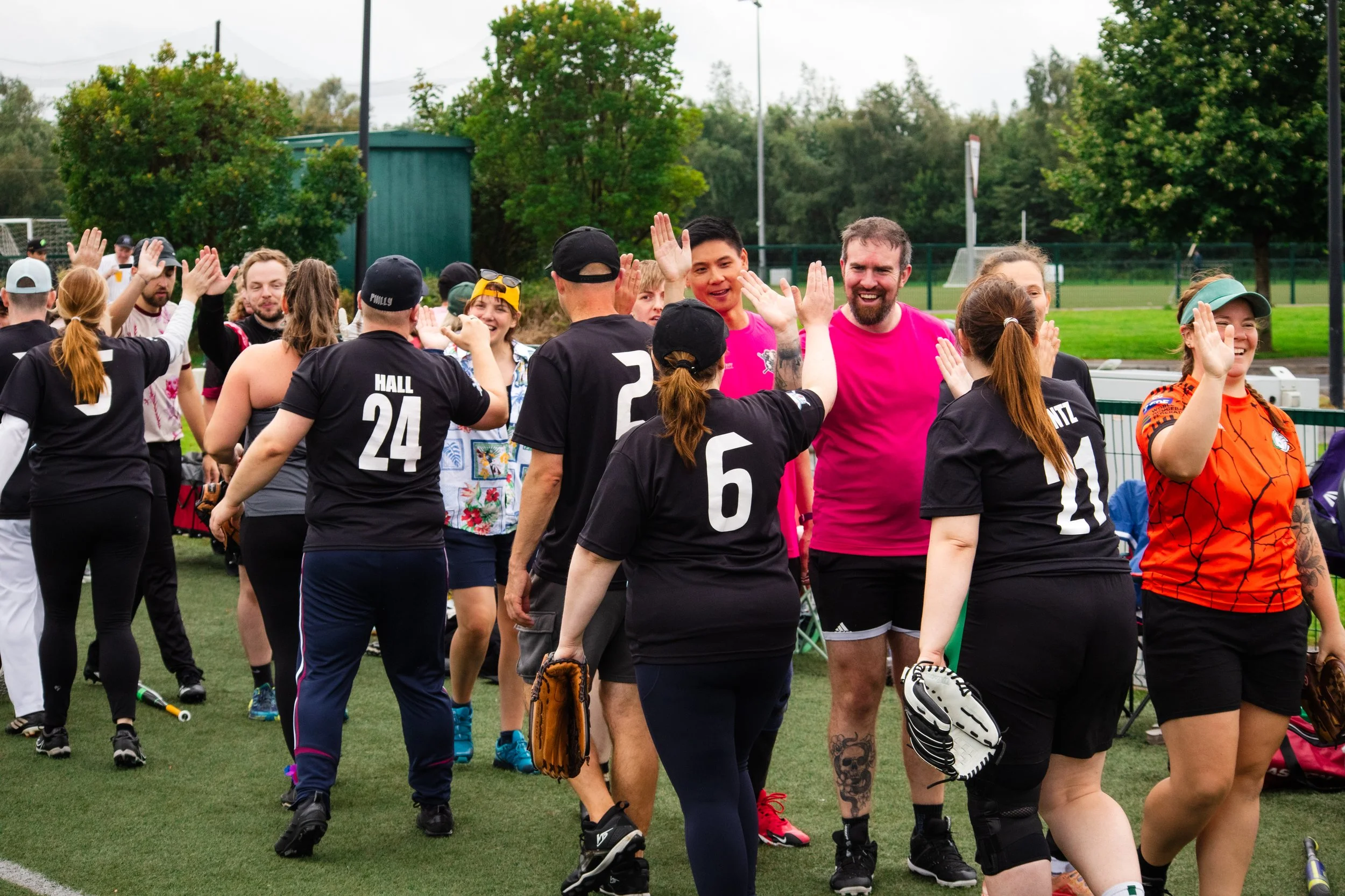 People at a sports field high-fiving and smiling after a game, with some wearing black and pink sportswear, some holding baseball gloves.