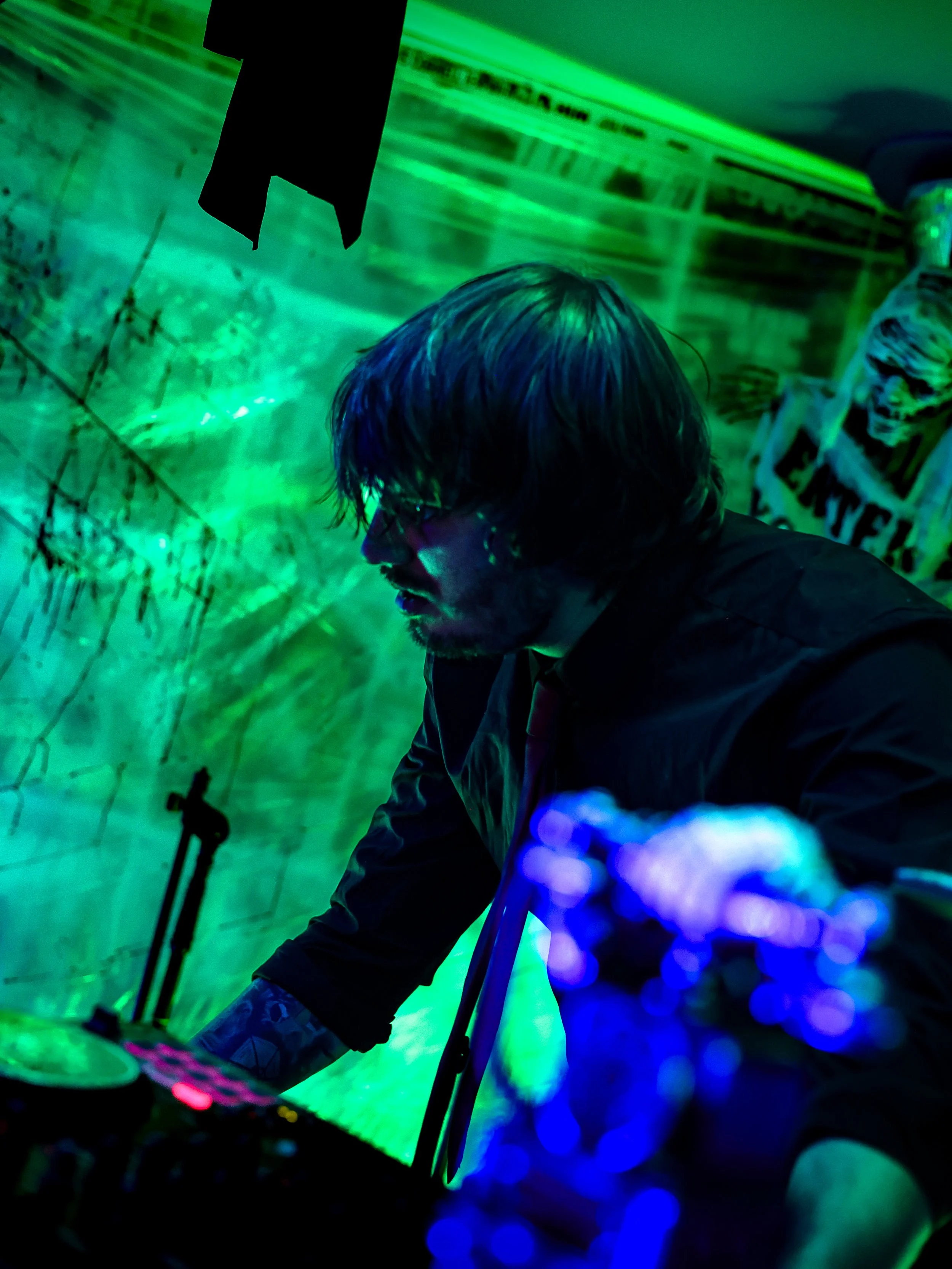 A man with shoulder-length hair and glasses working at a DJ station in a dimly lit room with green and blue lights.