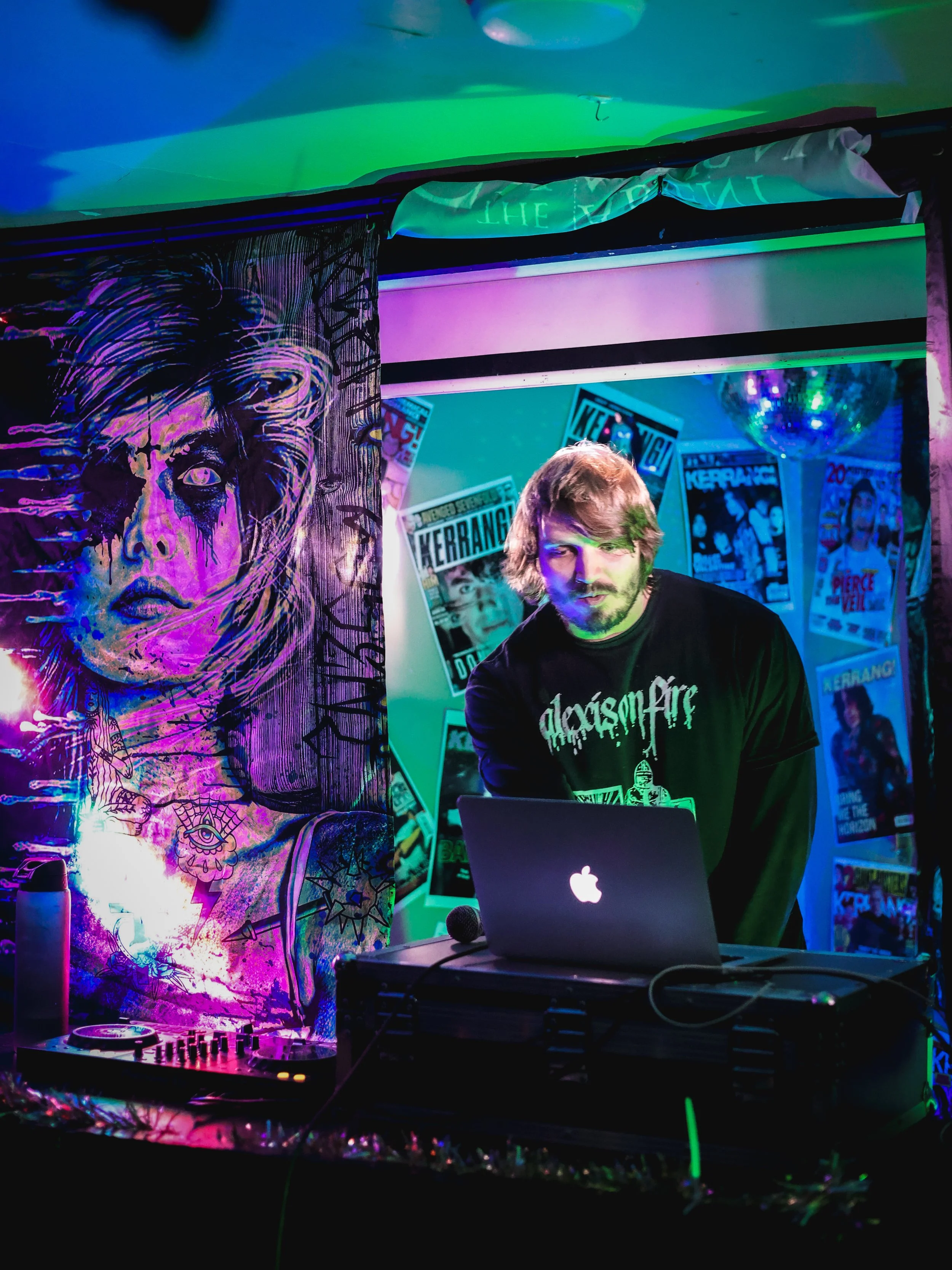 A DJ with shoulder-length hair and a beard is performing at a colorful, neon-lit venue. Behind him is a wall decorated with posters and artwork, including a large, purple and black illustration of a woman's face and eyes. There is a laptop, a microph