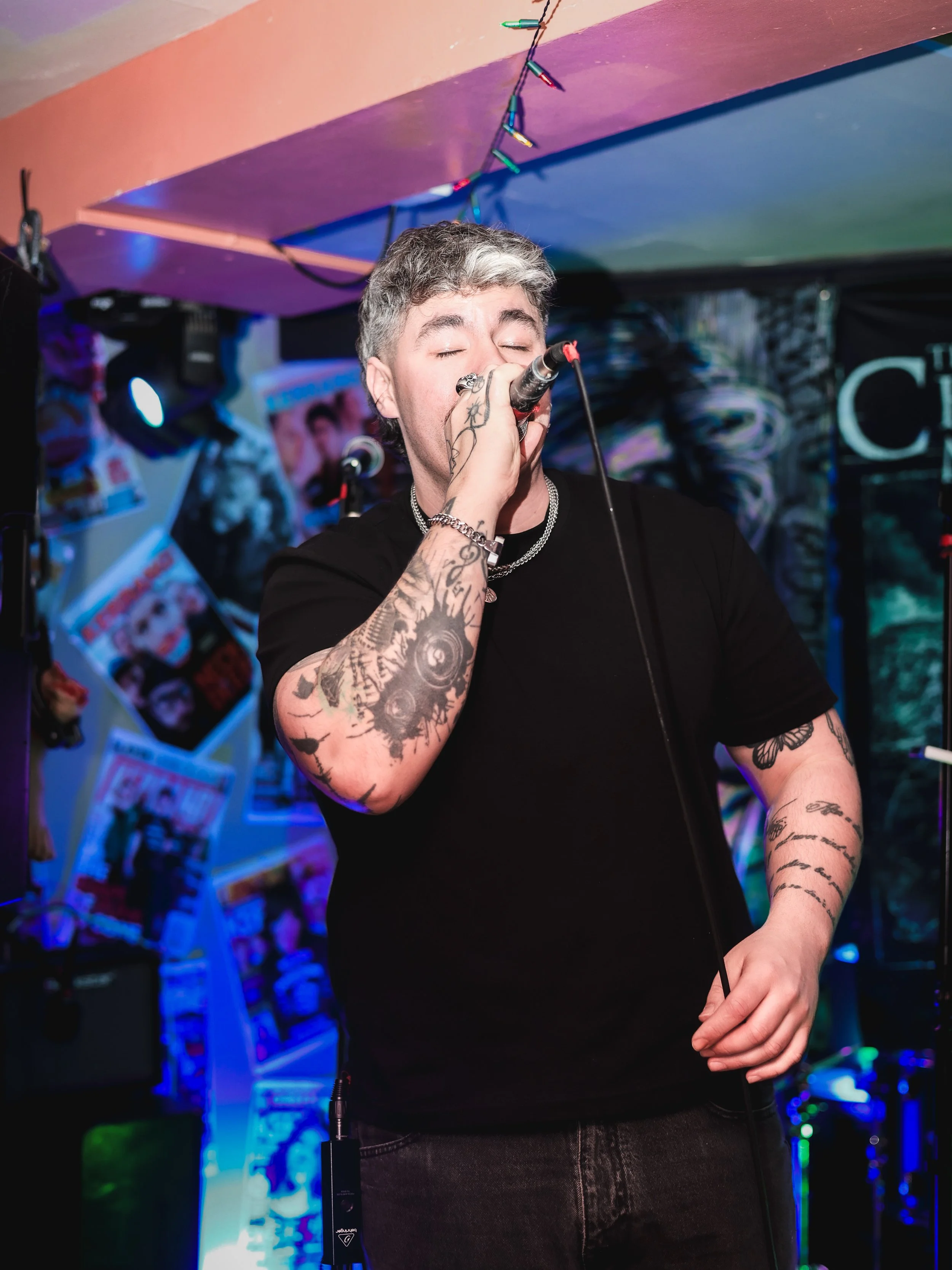 A man singing into a microphone at a music venue, with colorful posters and magazines on the wall behind him, and string lights hanging above.