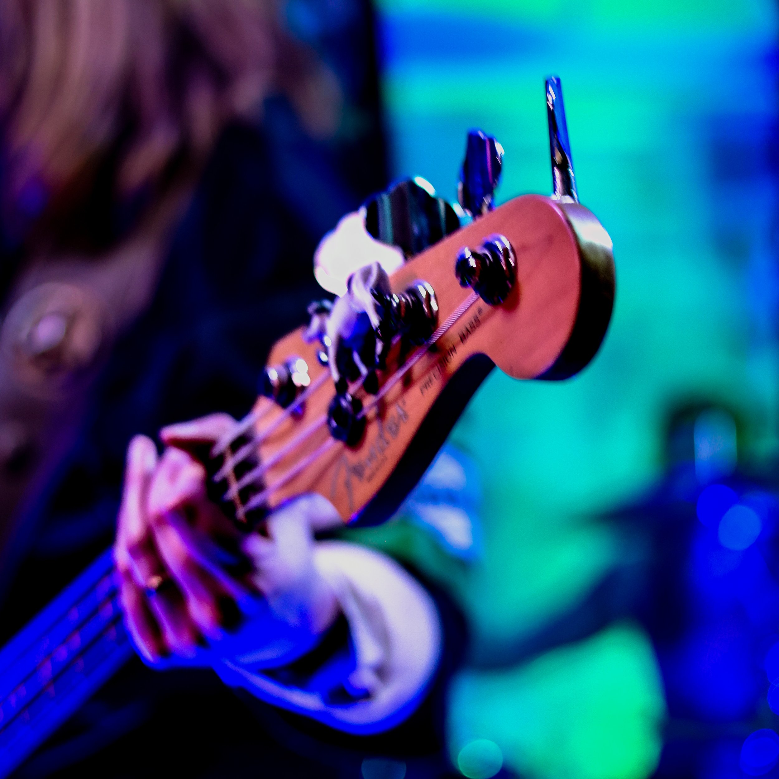 Close-up of a person playing an electric bass guitar on stage, with colorful stage lighting in the background.