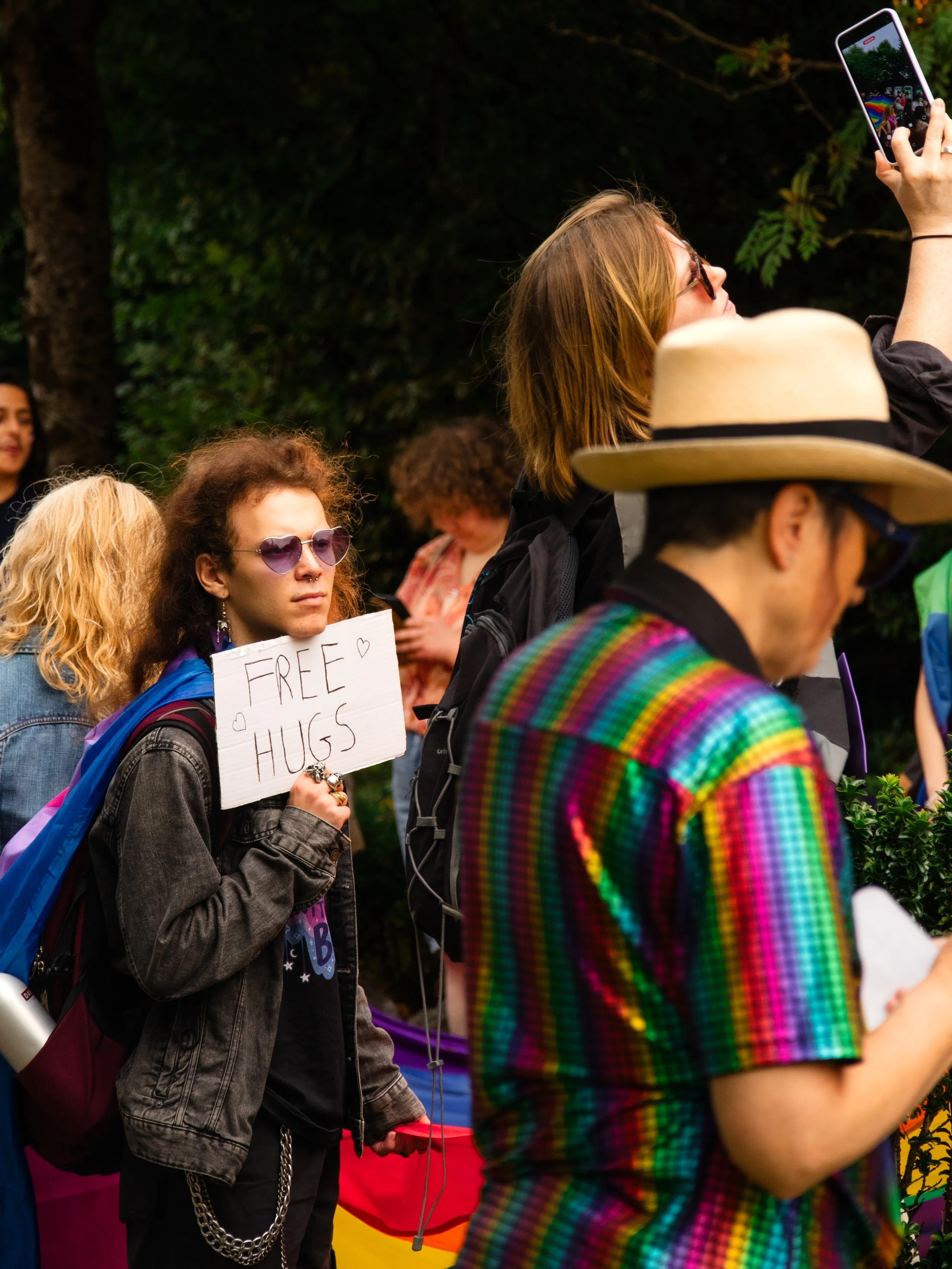 People at a gathering, with one person holding a sign that says 'FREE HUGS' and others taking photos, outdoors among trees.