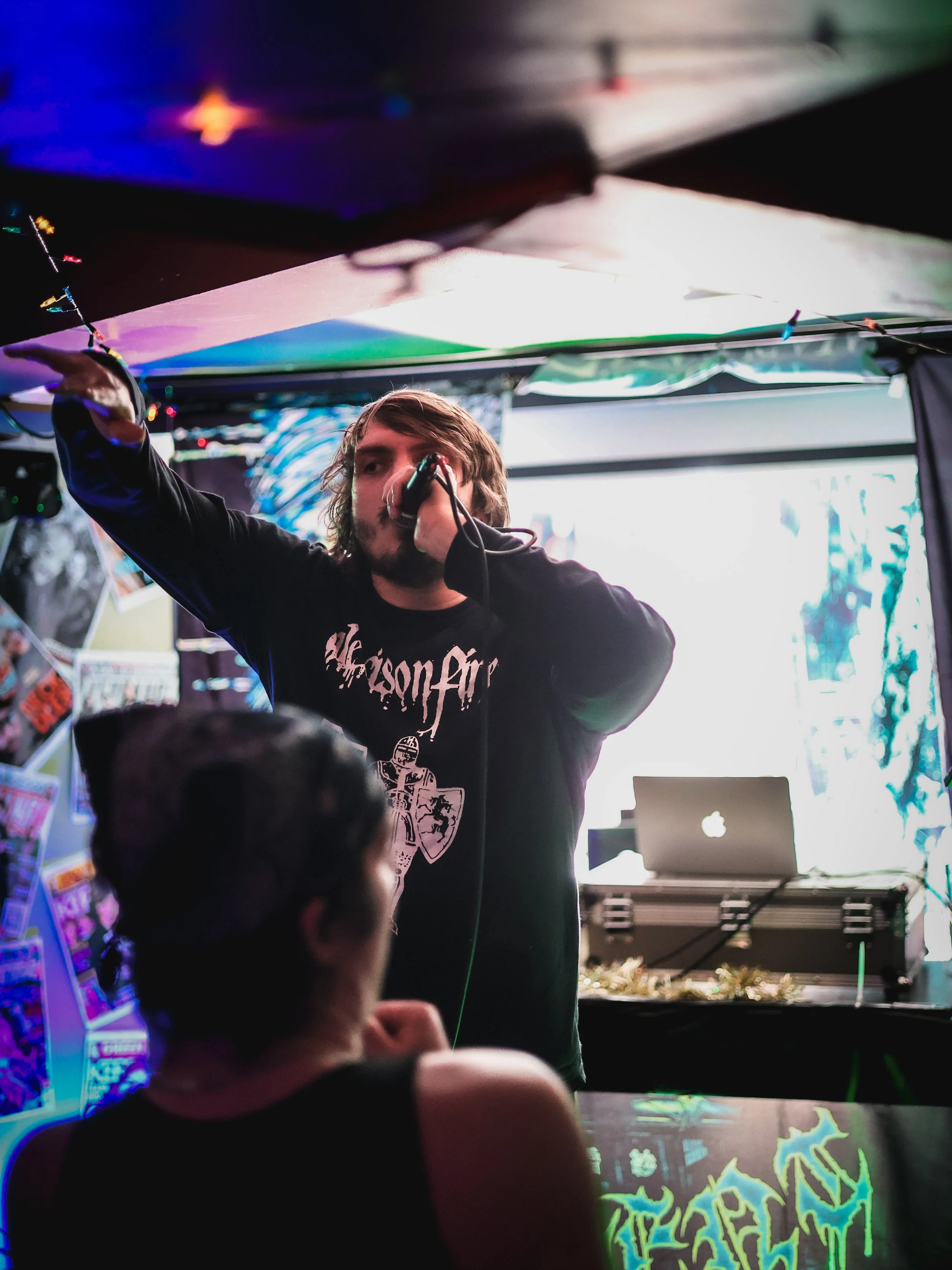 A young man with shoulder-length hair wearing a black album t-shirt is performing with a microphone in his right hand, on a stage with colorful lighting and posters on the wall behind him. A laptop and DJ equipment are on a table in the background, a
