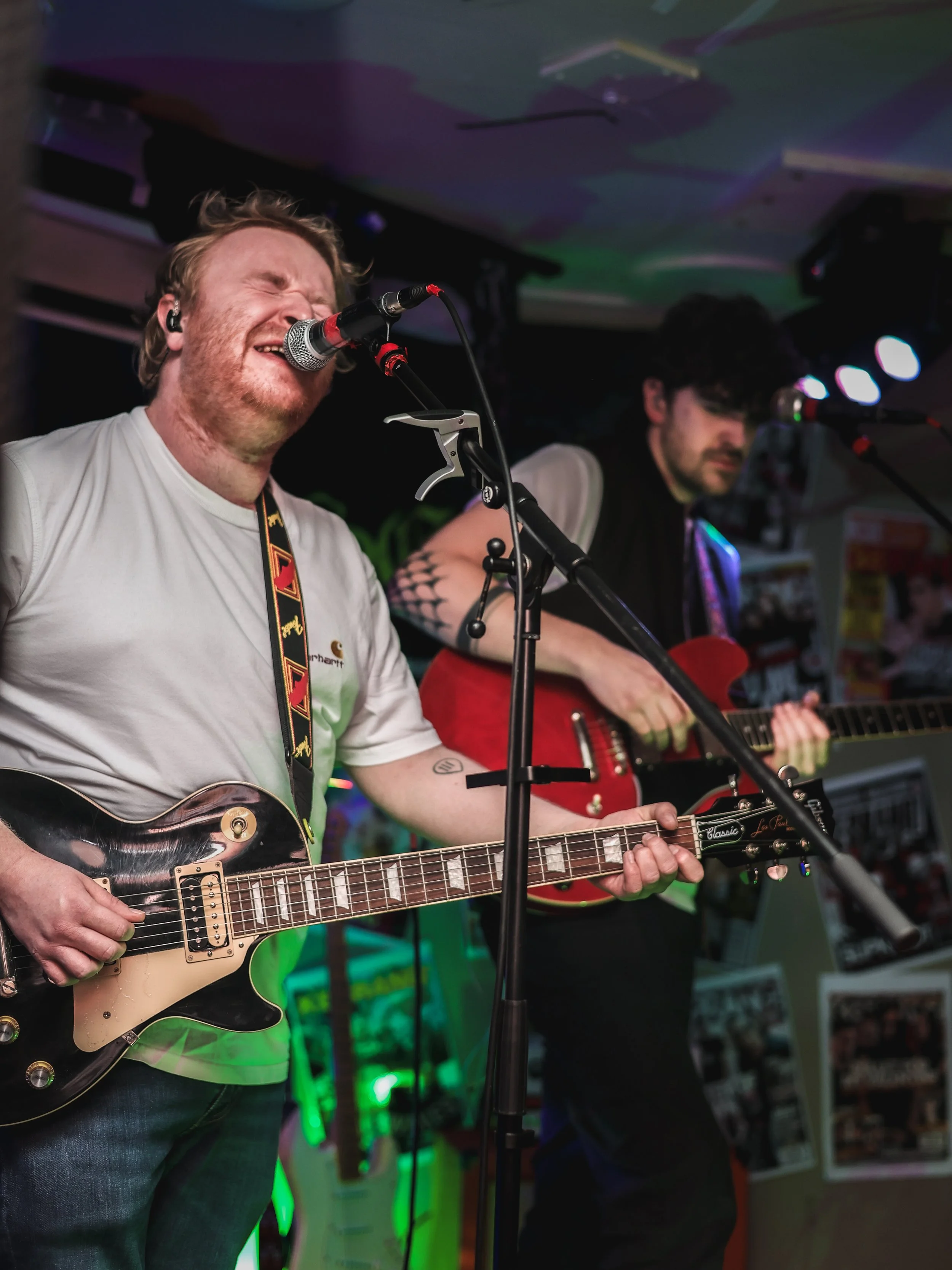 Two musicians perform on stage, playing guitars and singing into microphones, with one singing passionately with eyes closed.