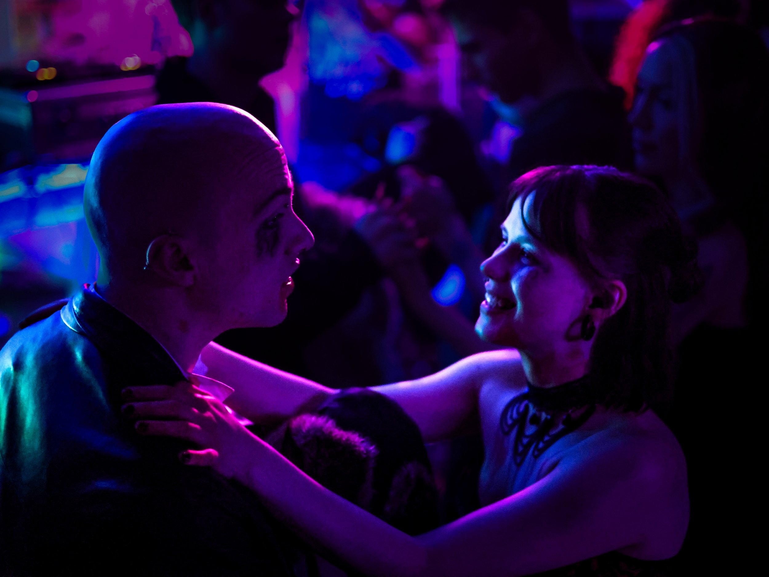 Two people, a man with a shaved head and a woman with short hair, are dancing closely at a party or nightclub illuminated by colorful neon lights. They are smiling at each other, and the woman is touching the man's shoulder.