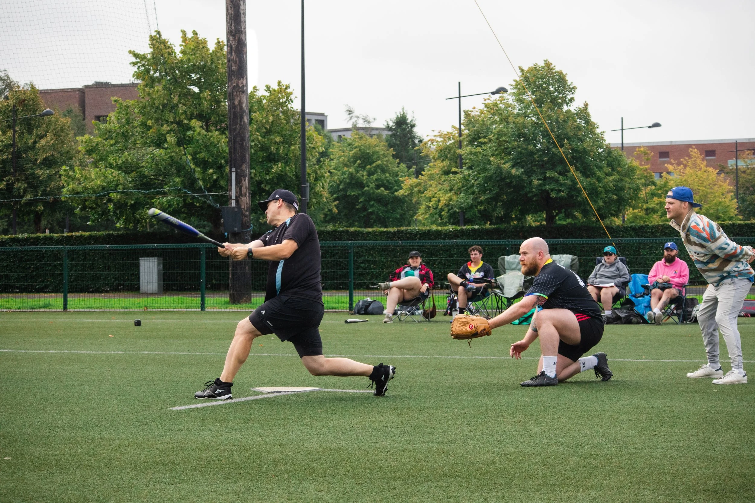 Group of people playing baseball on a field; one player is batting, others are sitting and watching in the background.