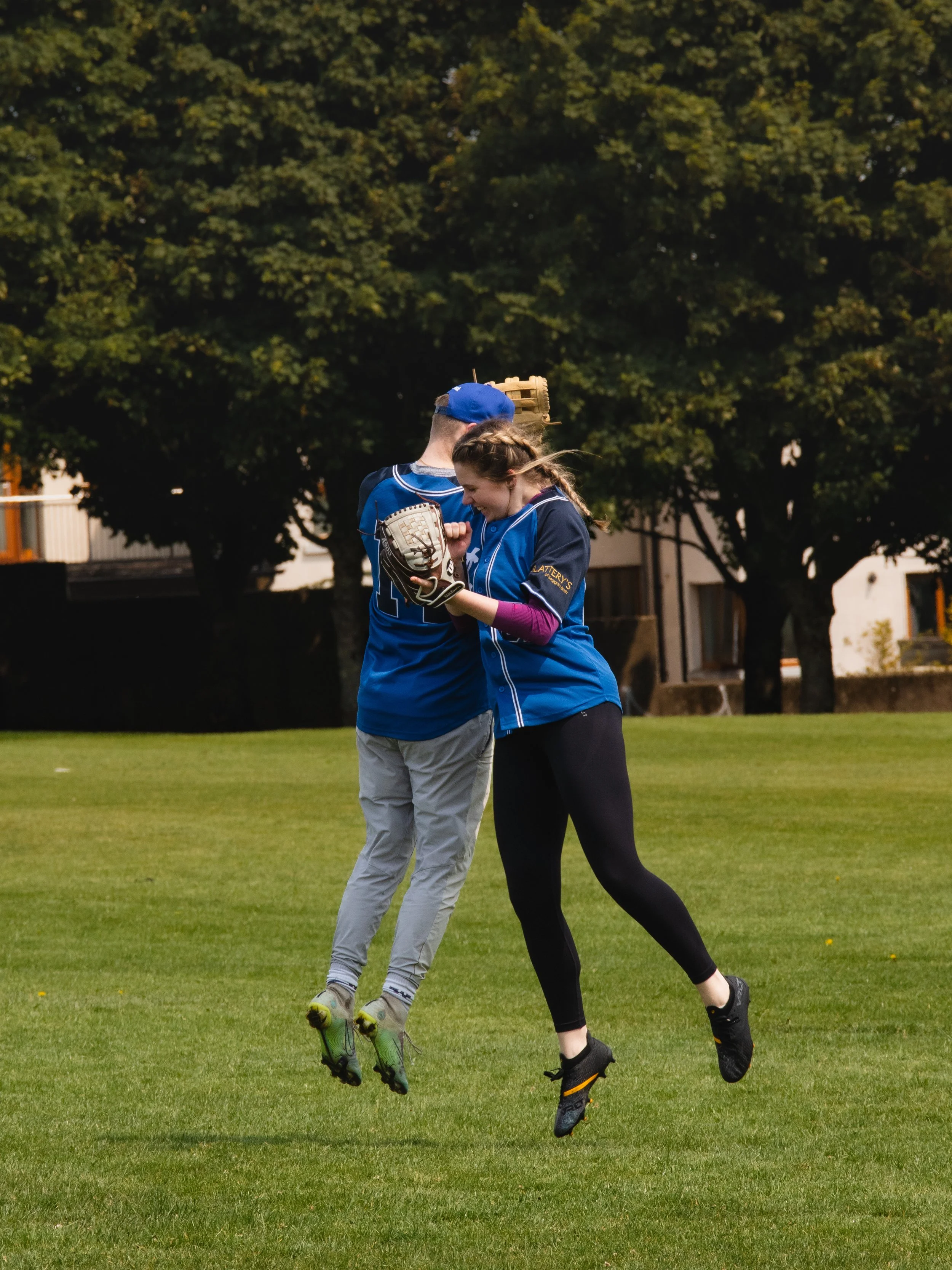 Two softball players in blue uniforms are jumping and celebrating on the field, clutching a baseball glove together with trees and buildings in the background.