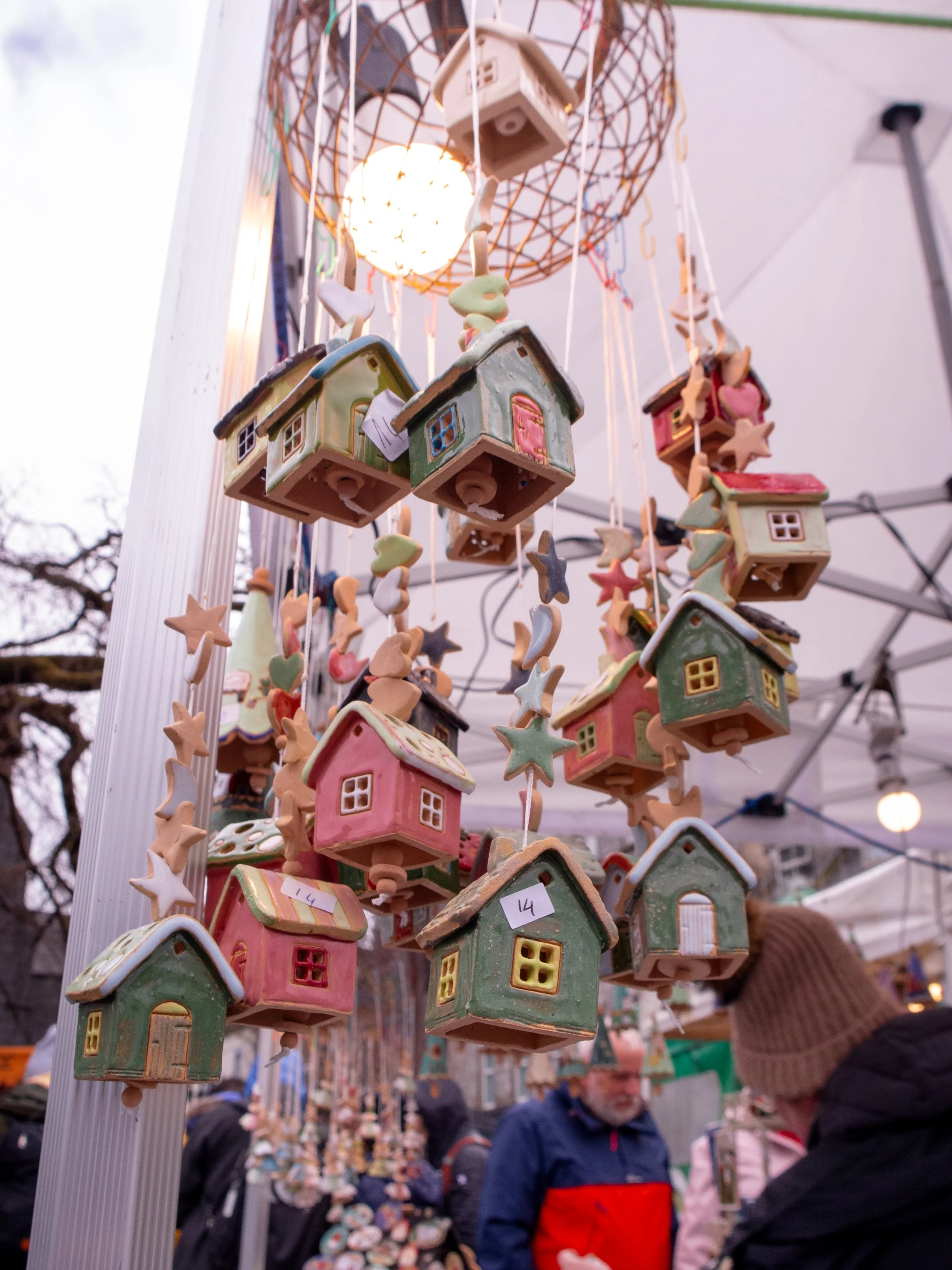 Colorful ceramic Christmas houses and ornaments hanging at a market stall, with people browsing in the background.