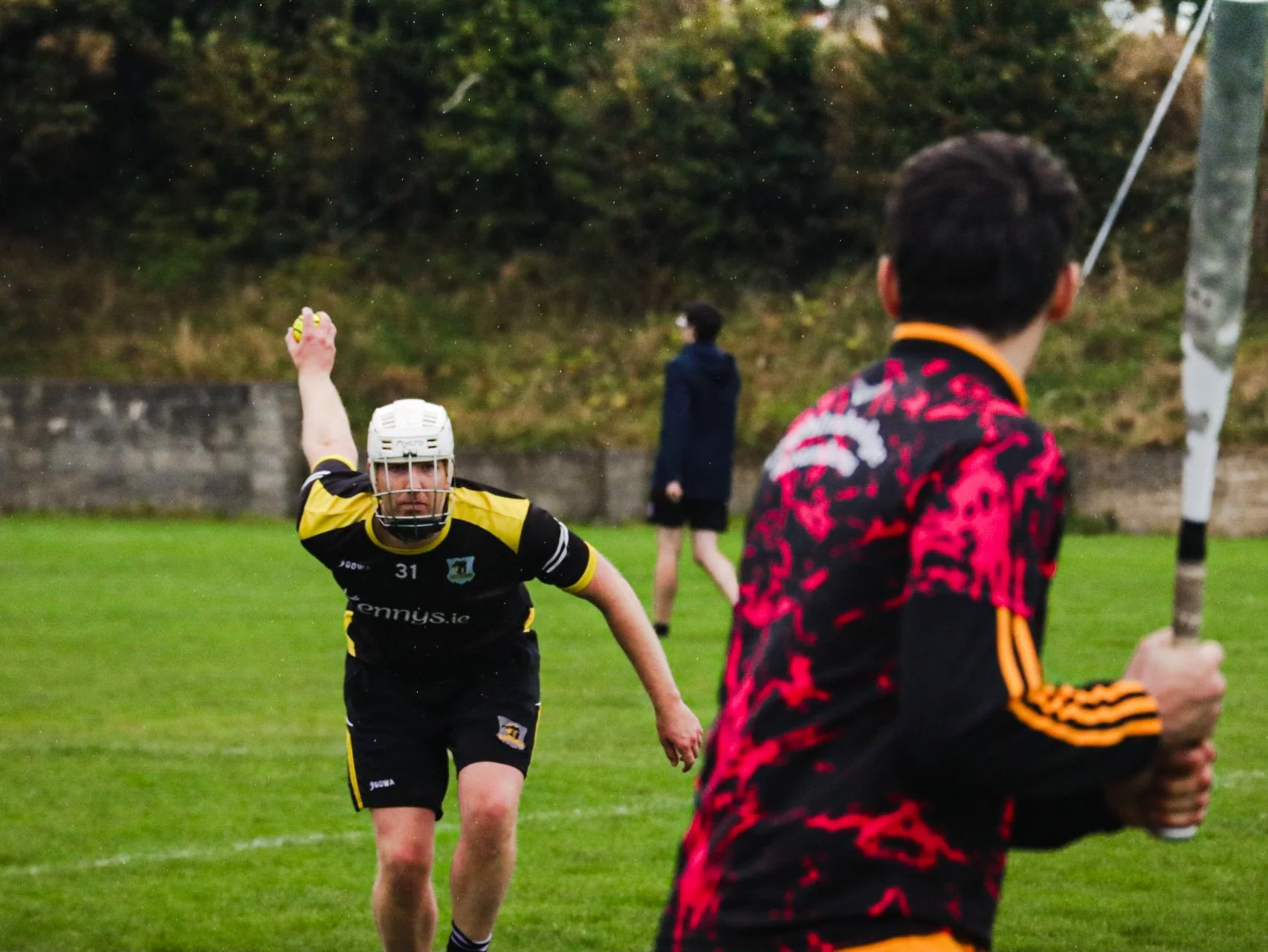 A camogie player in a yellow and black uniform prepares to catch a ball, while another player in a black and red uniform holds a hurley in the foreground on a grassy field.
