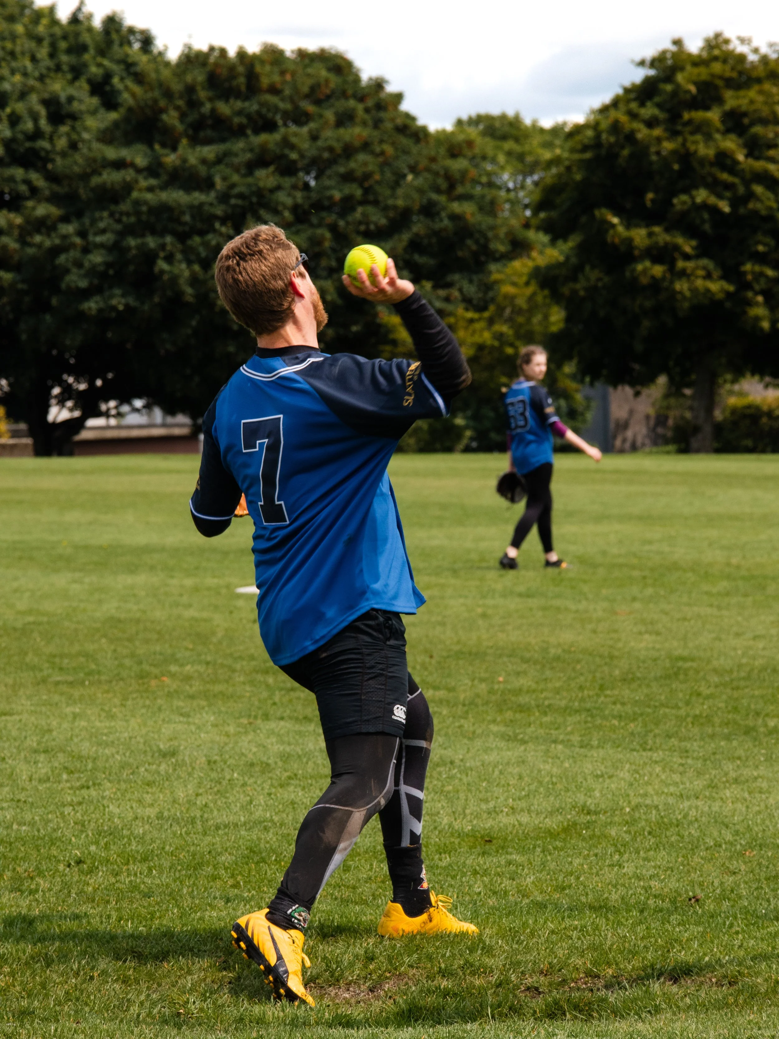 A person playing softball on a grassy field, preparing to throw a bright yellow ball, with another person in the background and trees around.