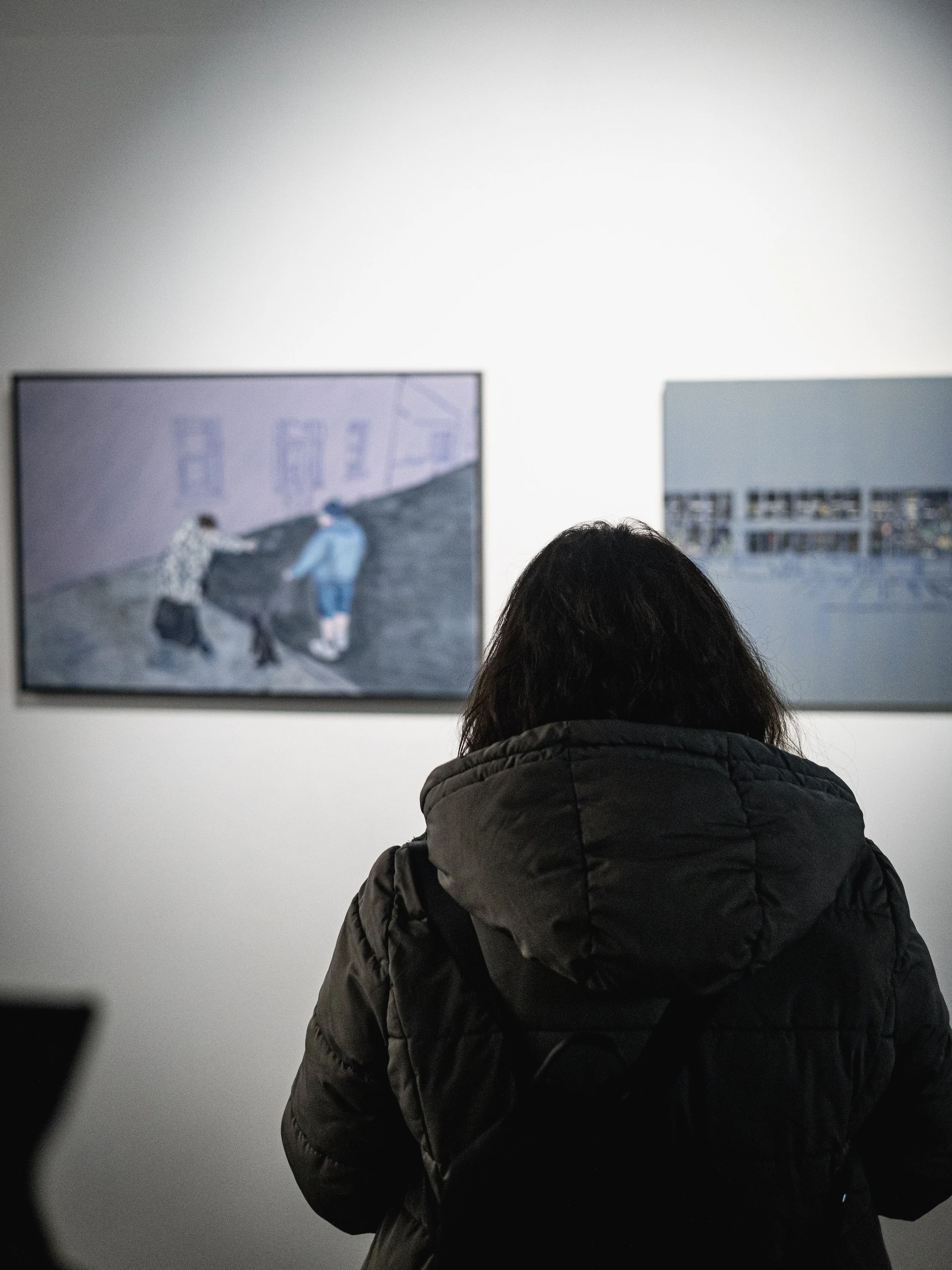 A person wearing a black jacket with a hood standing in front of a white wall, looking at two framed artworks in a gallery.