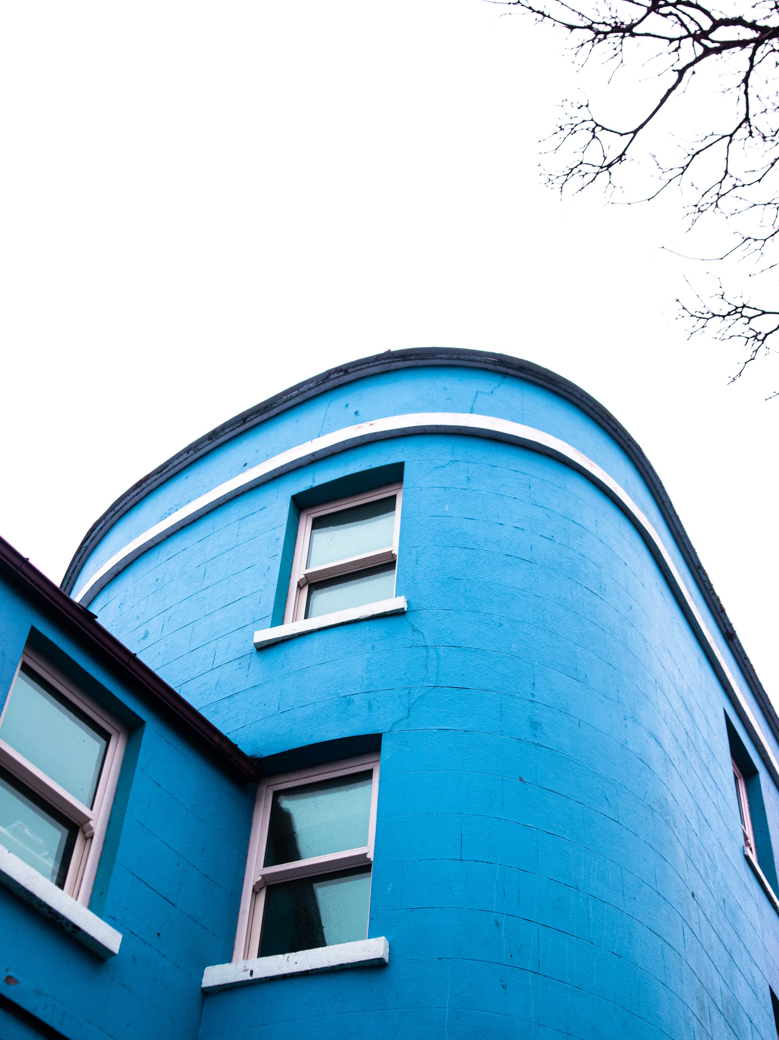 A bright blue building with three windows, showing a rounded corner design, against an overcast sky with bare tree branches.