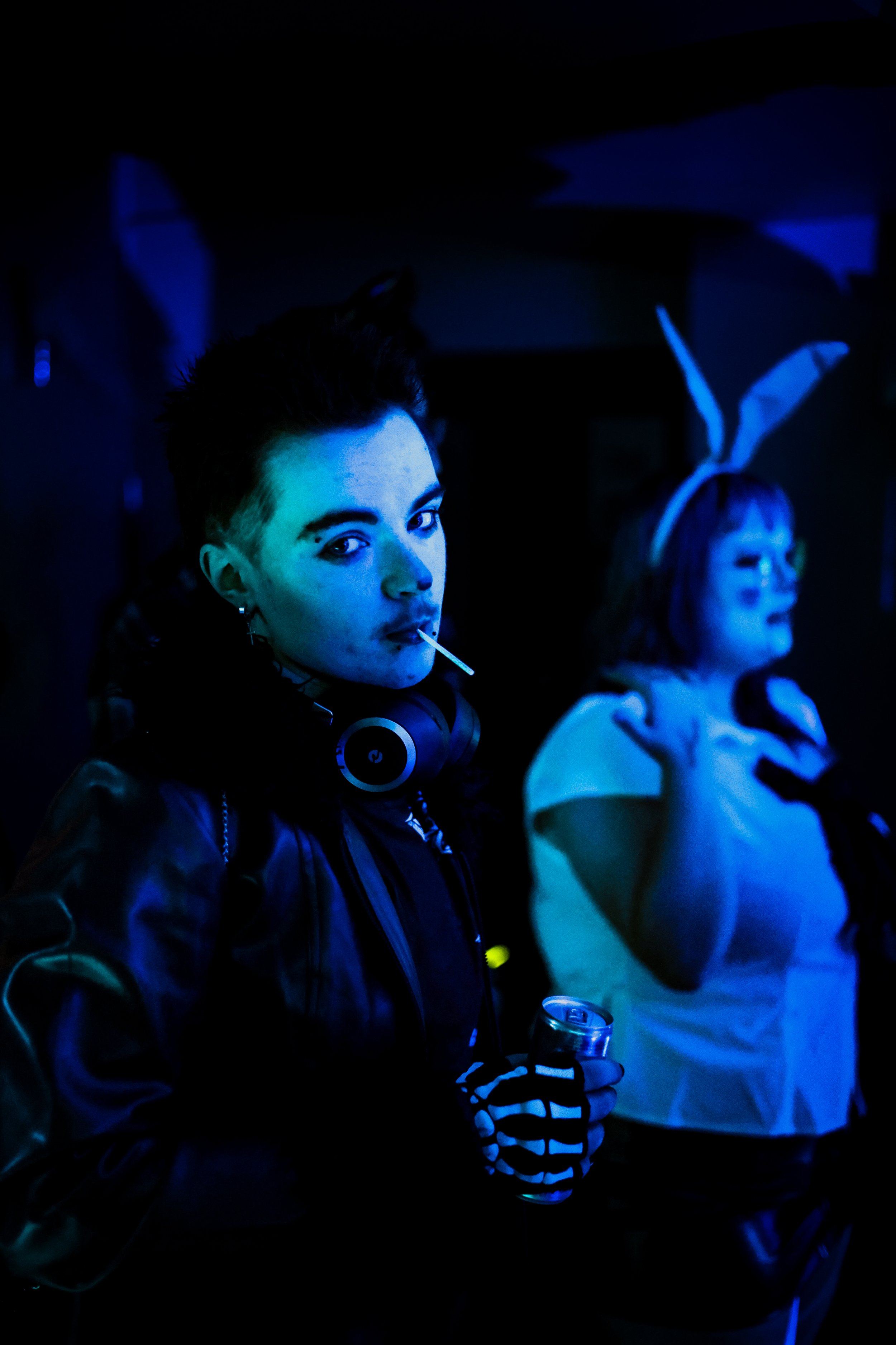 A person with short dark hair, wearing a leather jacket and striped gloves, is holding a can and looking at the camera with a lollipop in their mouth and headphones around their neck. Another person in the background is dressed in a costume with bunn