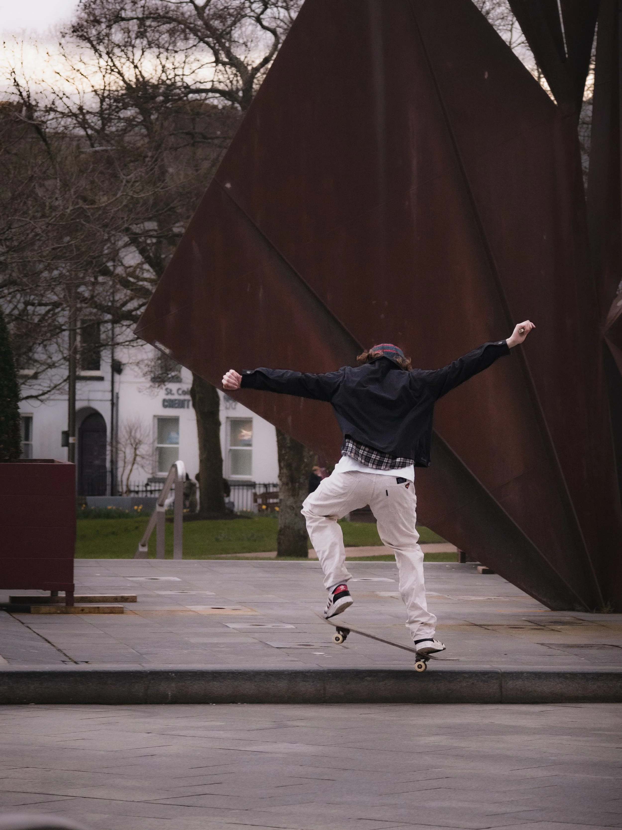 A skateboarder performing a trick on a sidewalk near a large abstract metal sculpture, with trees and buildings in the background during twilight.