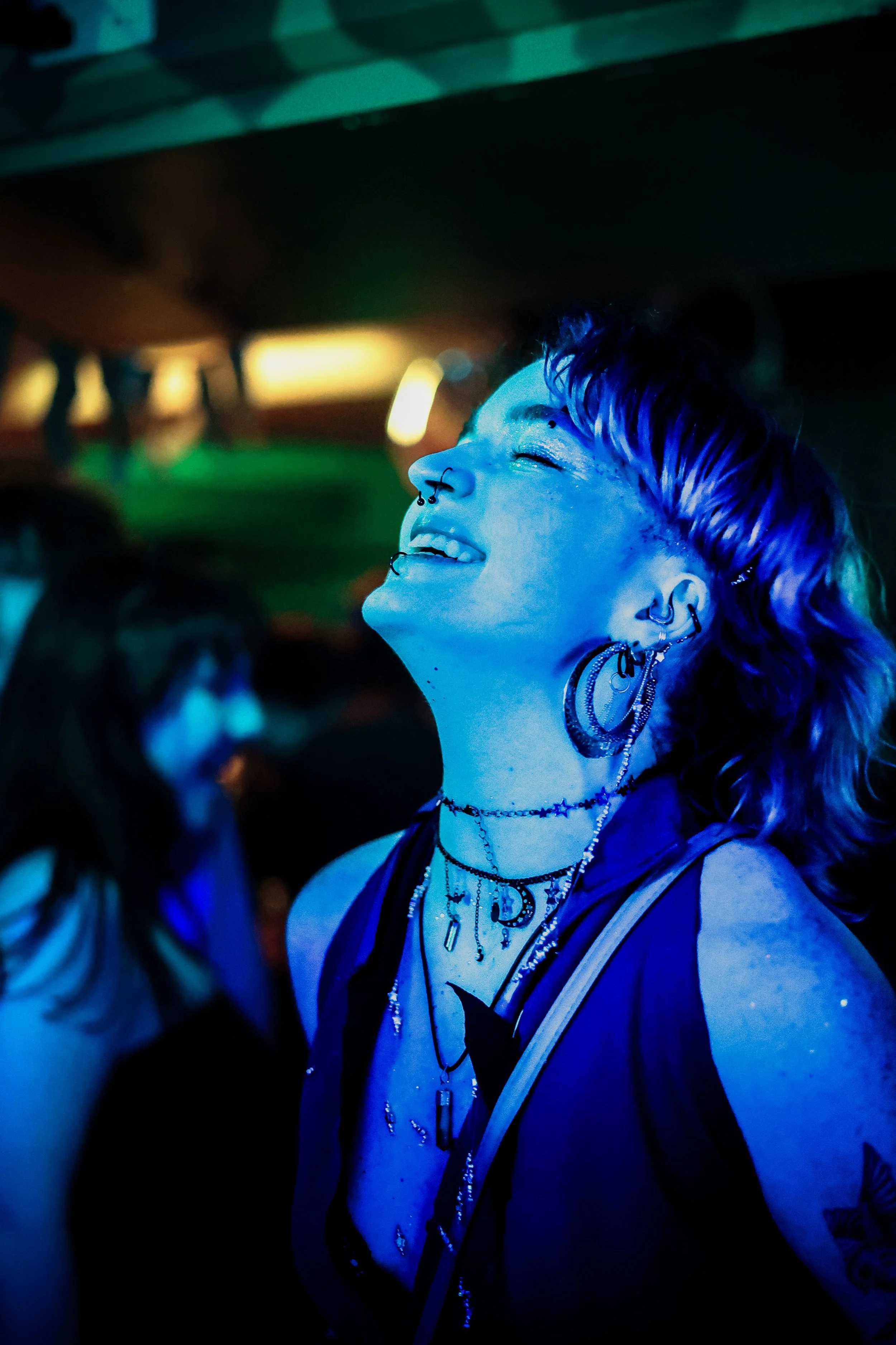 A young person with tattoos and piercings, smiling with their eyes closed, illuminated by blue lighting at a crowded event or party.