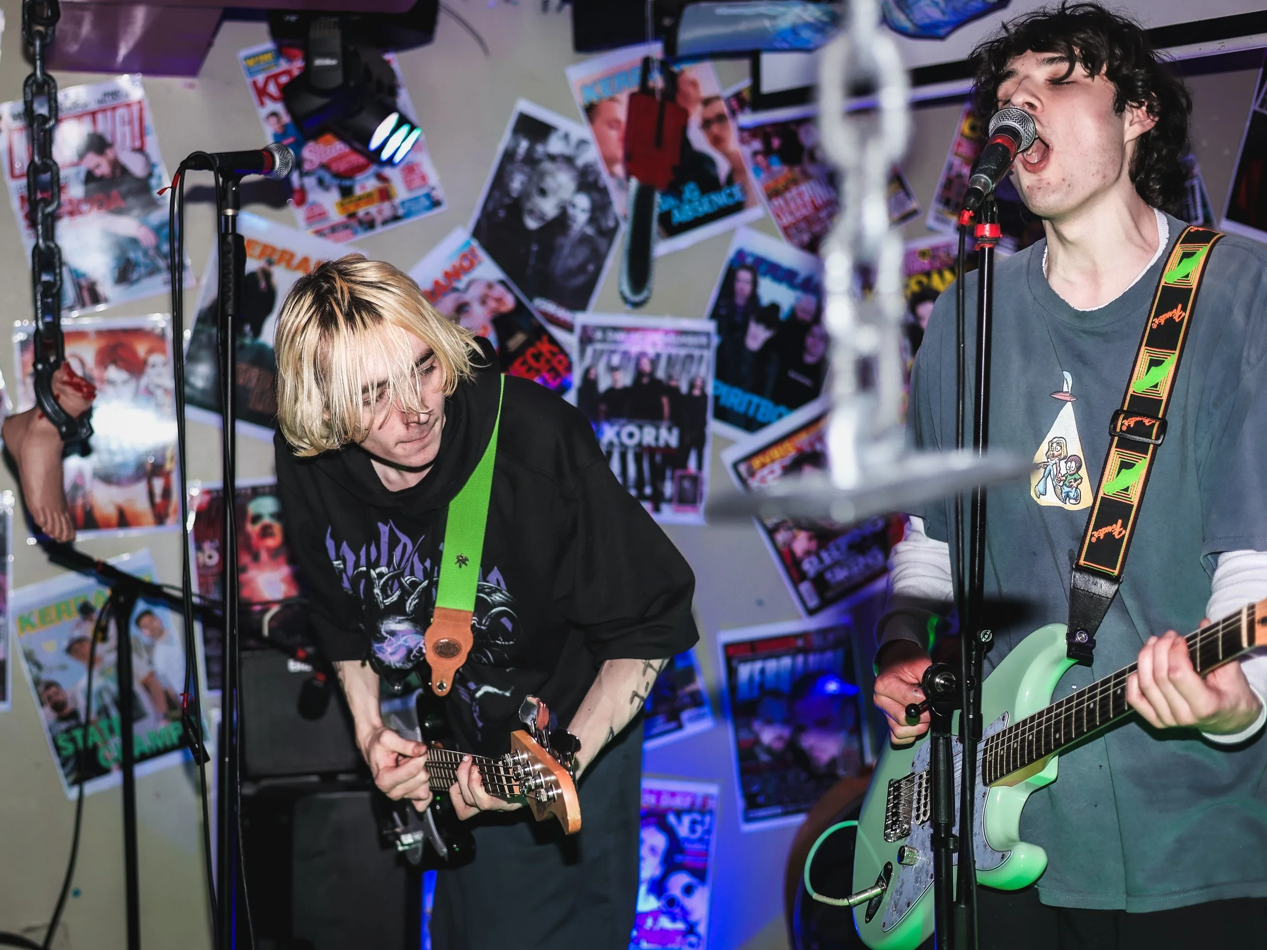 Two young men performing a live music set, one playing an electric guitar and the other singing into a microphone, with magazines and posters decorating the background.