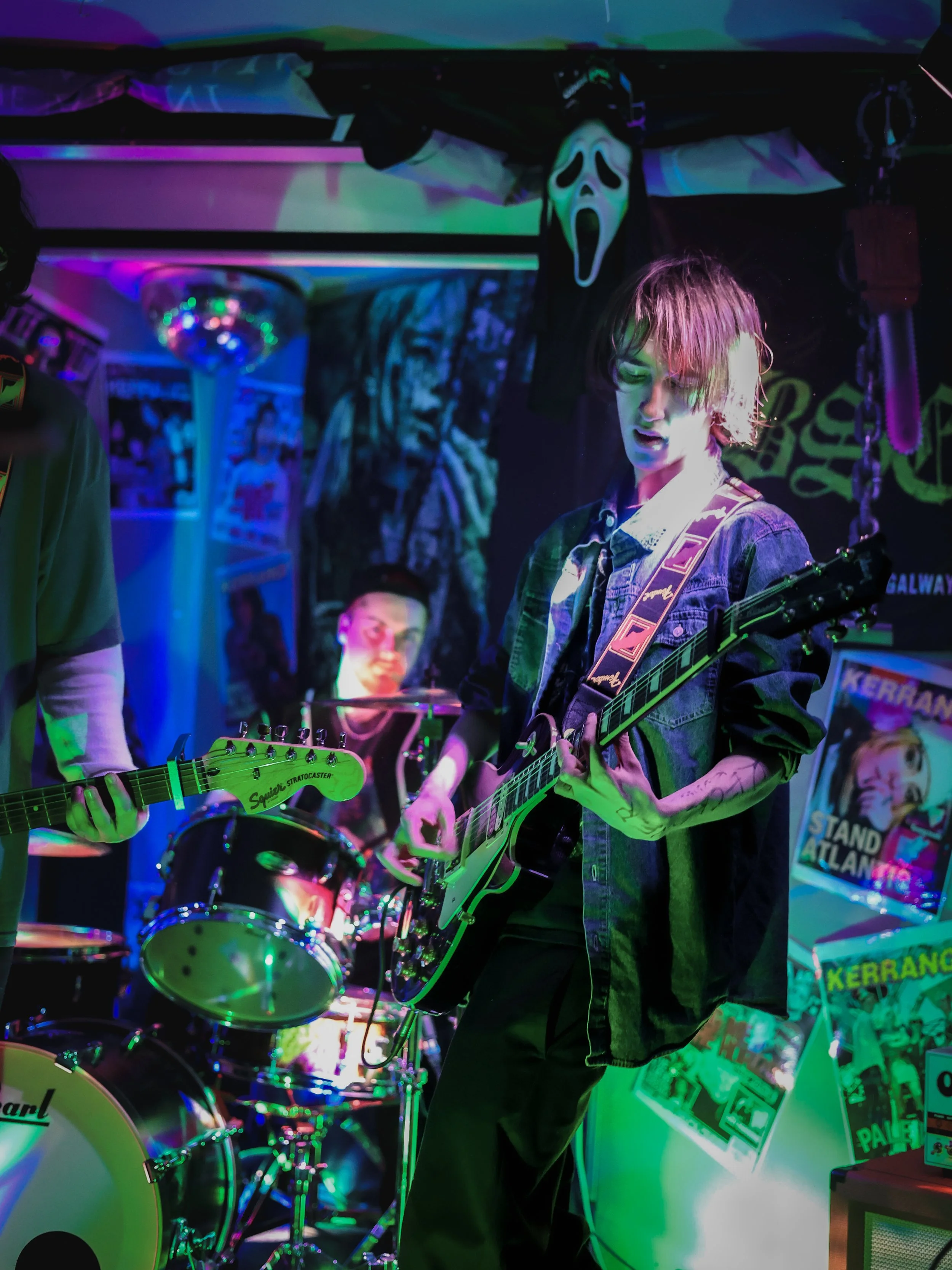Young person playing electric guitar in a band with a drummer in the background, neon lights, posters, and a creepy ghost face mask hanging from the ceiling, creating a vibrant, alternative music scene atmosphere.