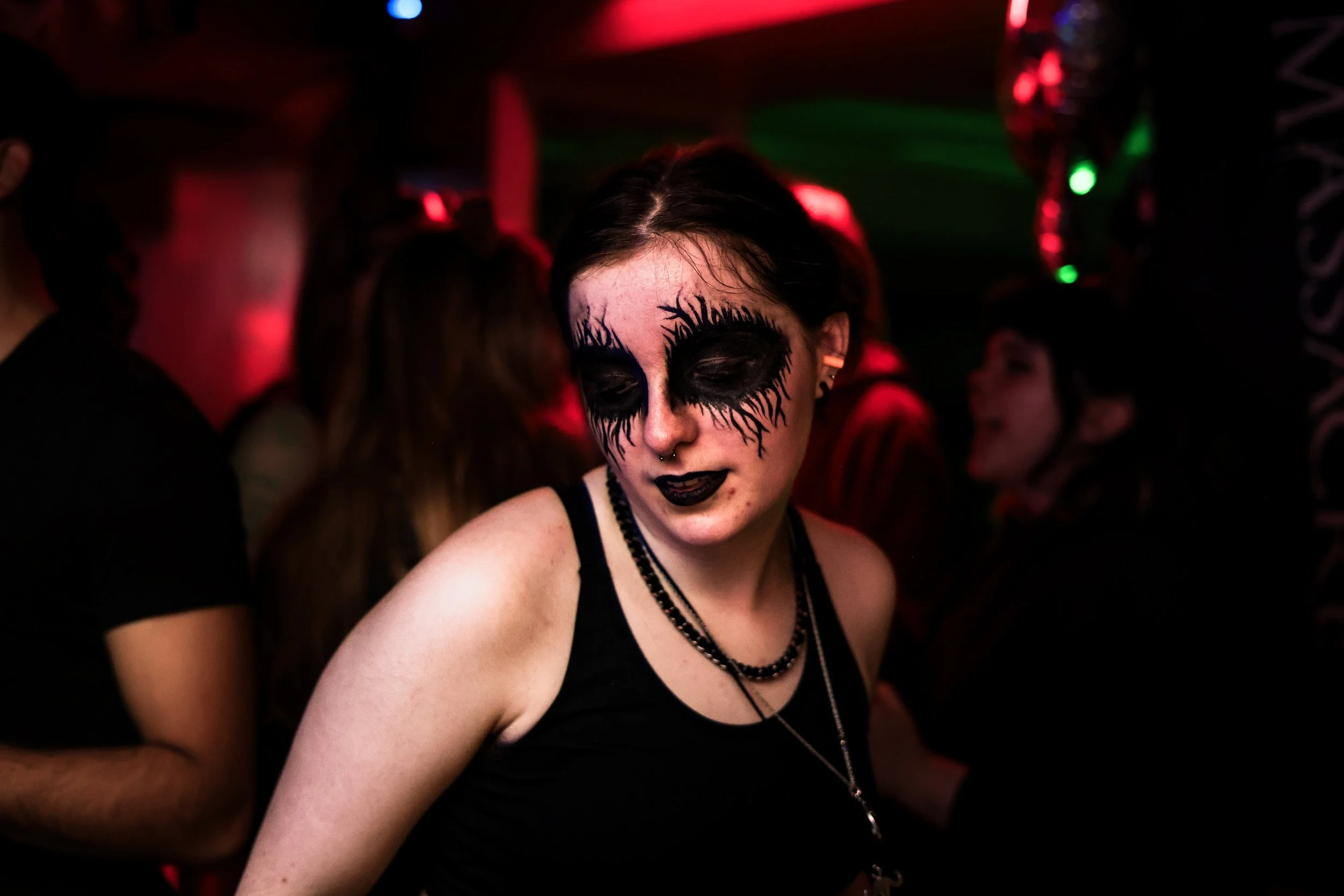 A woman with black makeup resembling dark, dripping eyeshadow around her eyes, black lipstick, and a septum piercing, seen at a dark, red-lit party or nightclub.
