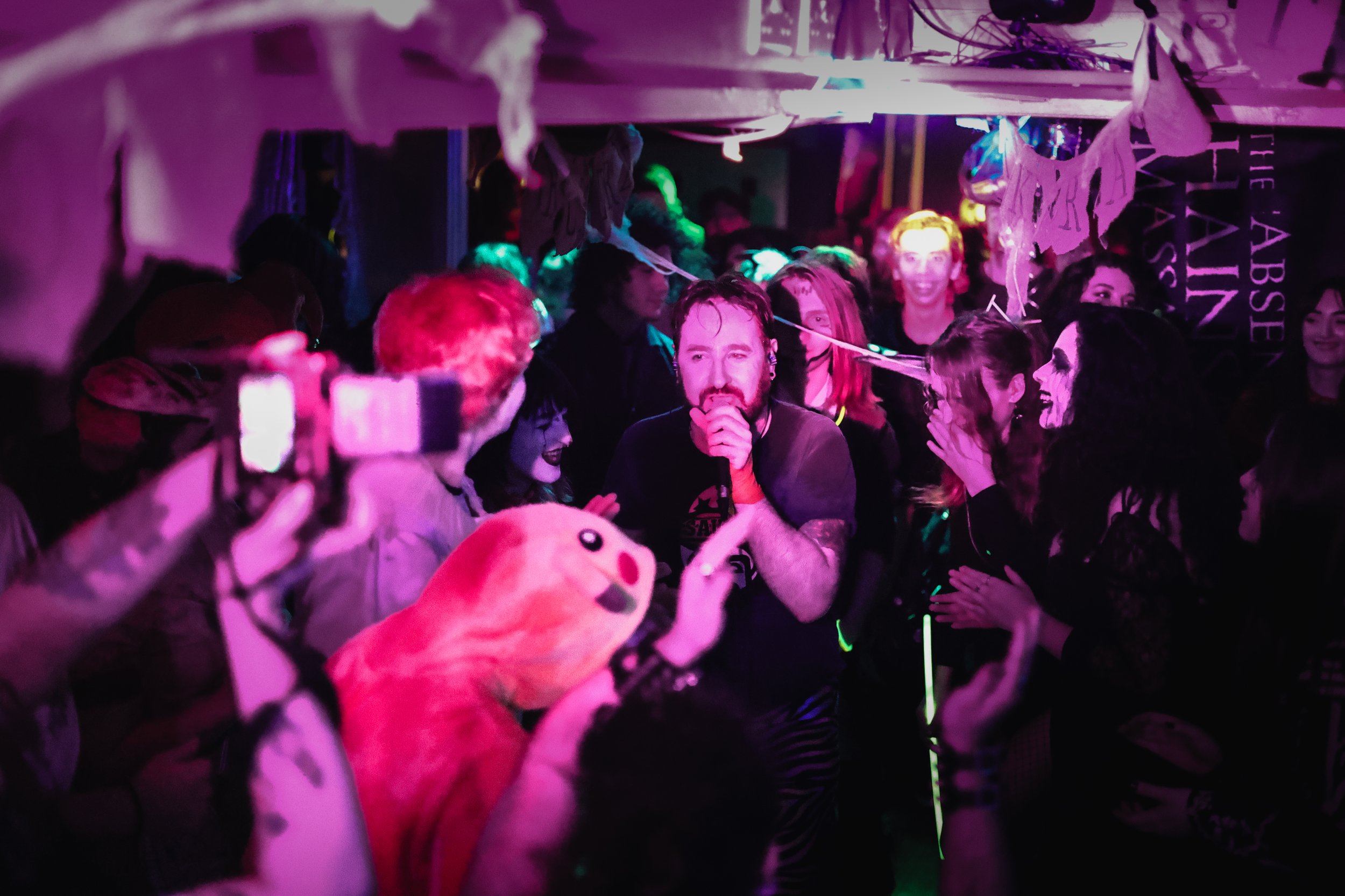 A man with a microphone singing or speaking in front of a crowd at a party or event. The crowd is lively, with some people taking photos, others clapping, and some wearing costumes or accessories. The setting has neon and colored lighting, creating a
