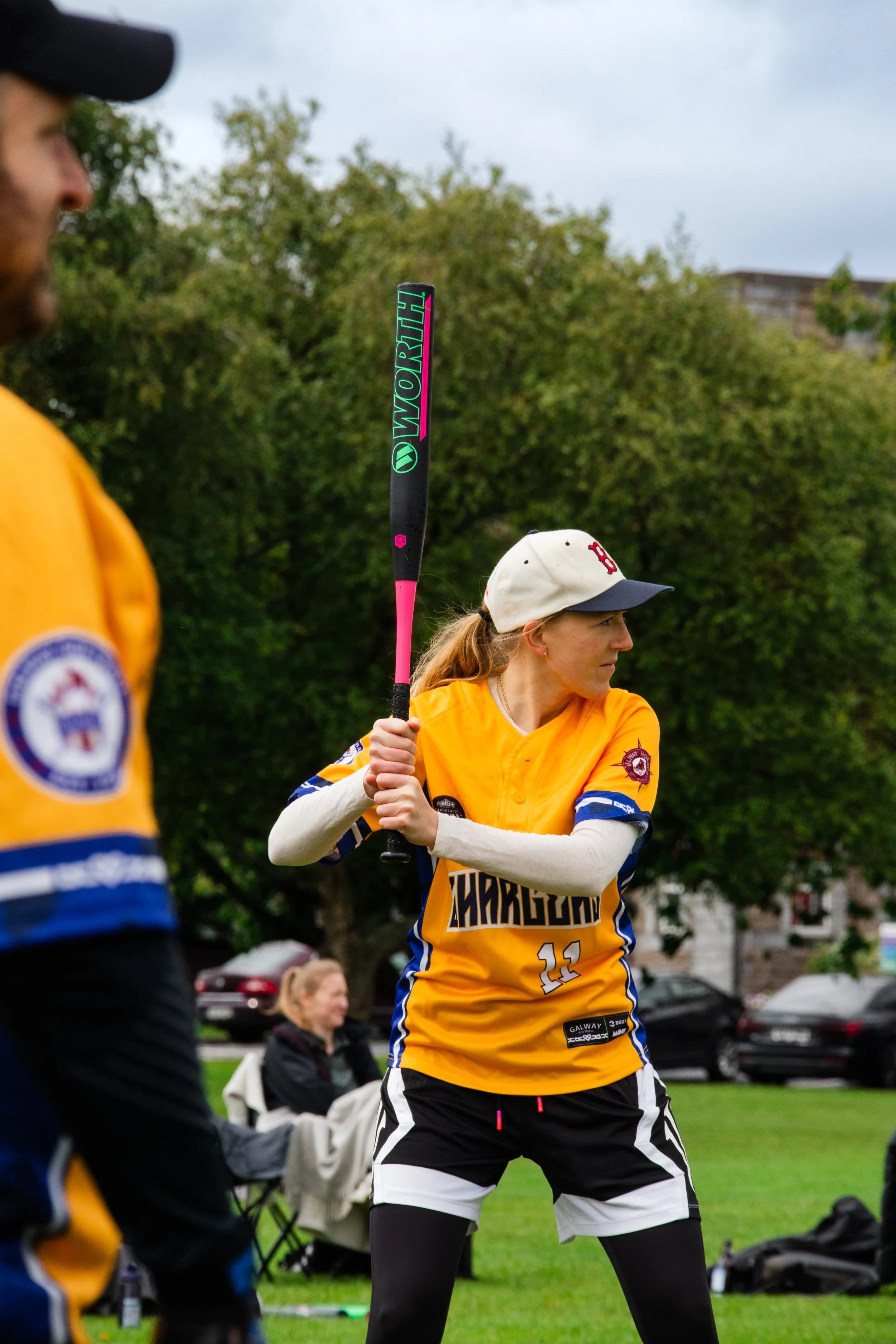 A woman wearing a yellow sports jersey, black and white shorts, black leggings, and a white baseball cap is holding a black and pink softball bat, preparing to swing. Another person in a similar jersey is visible partially on the left, and a seated w