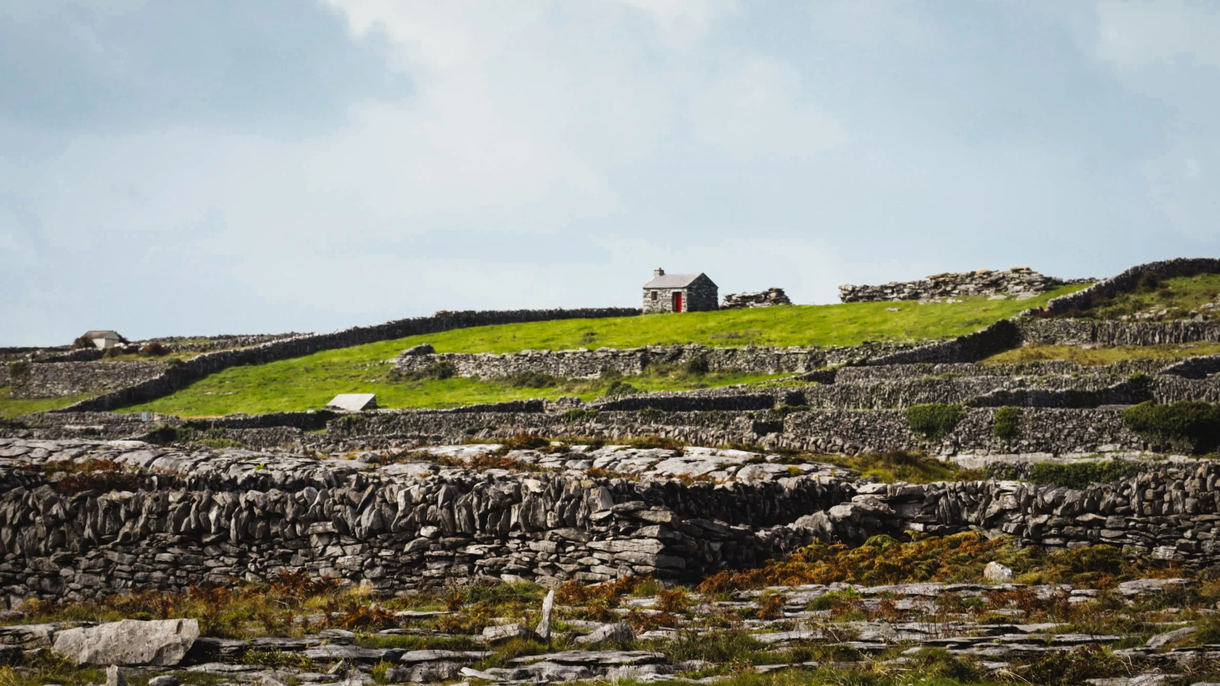 A rural landscape with layered stone walls and small houses on green hills under a cloudy sky.