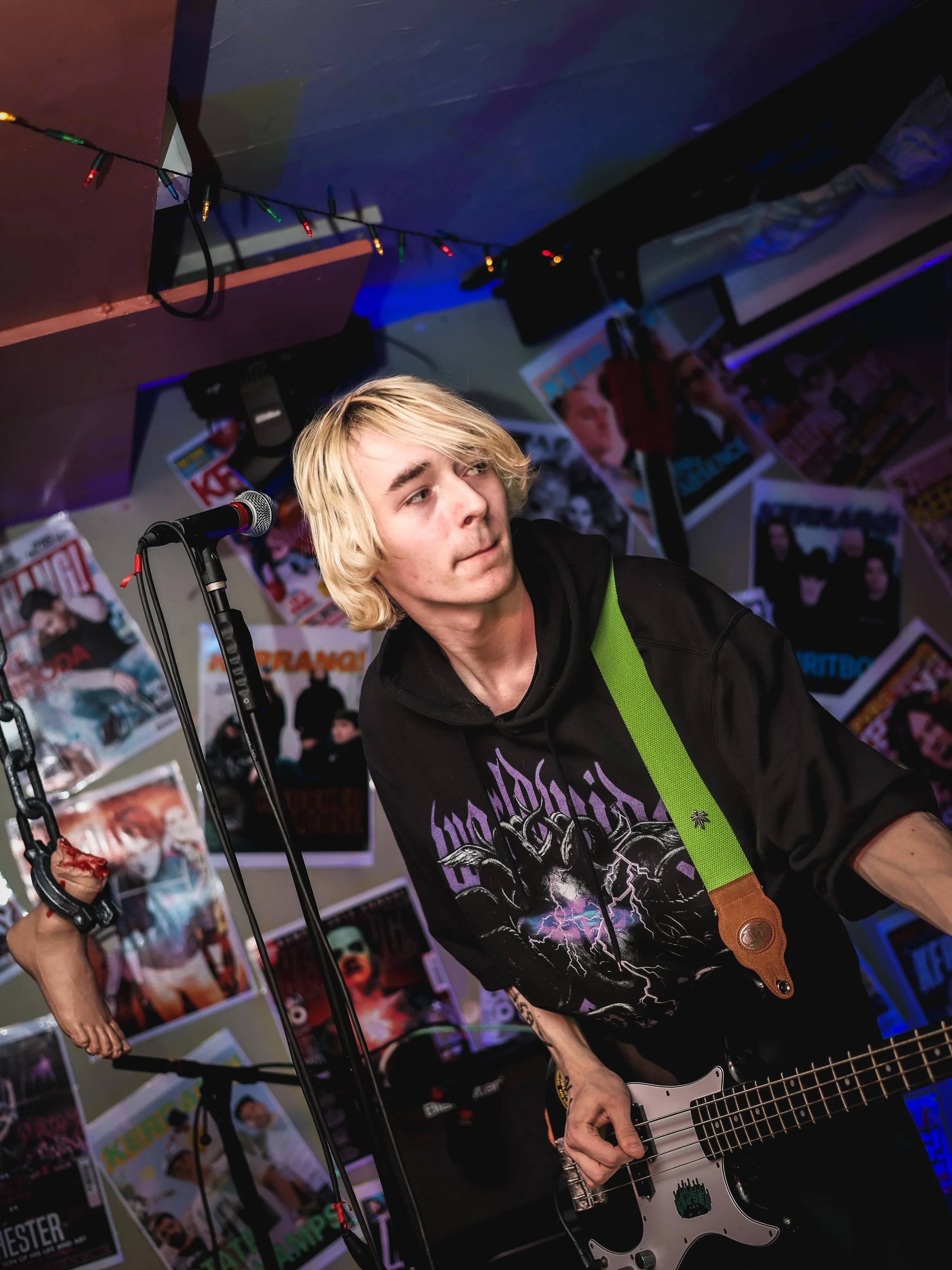 A young man with blond hair playing electric bass guitar in a music venue decorated with posters on the walls.