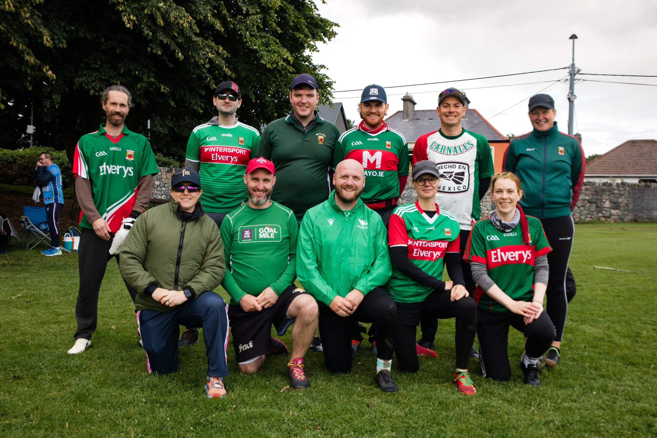 A group of eleven people, including men and women, wearing athletic gear and jerseys, posing for a photo outdoors on a grassy field with trees and houses in the background. They are smiling and kneeling or standing, likely after a sports event.