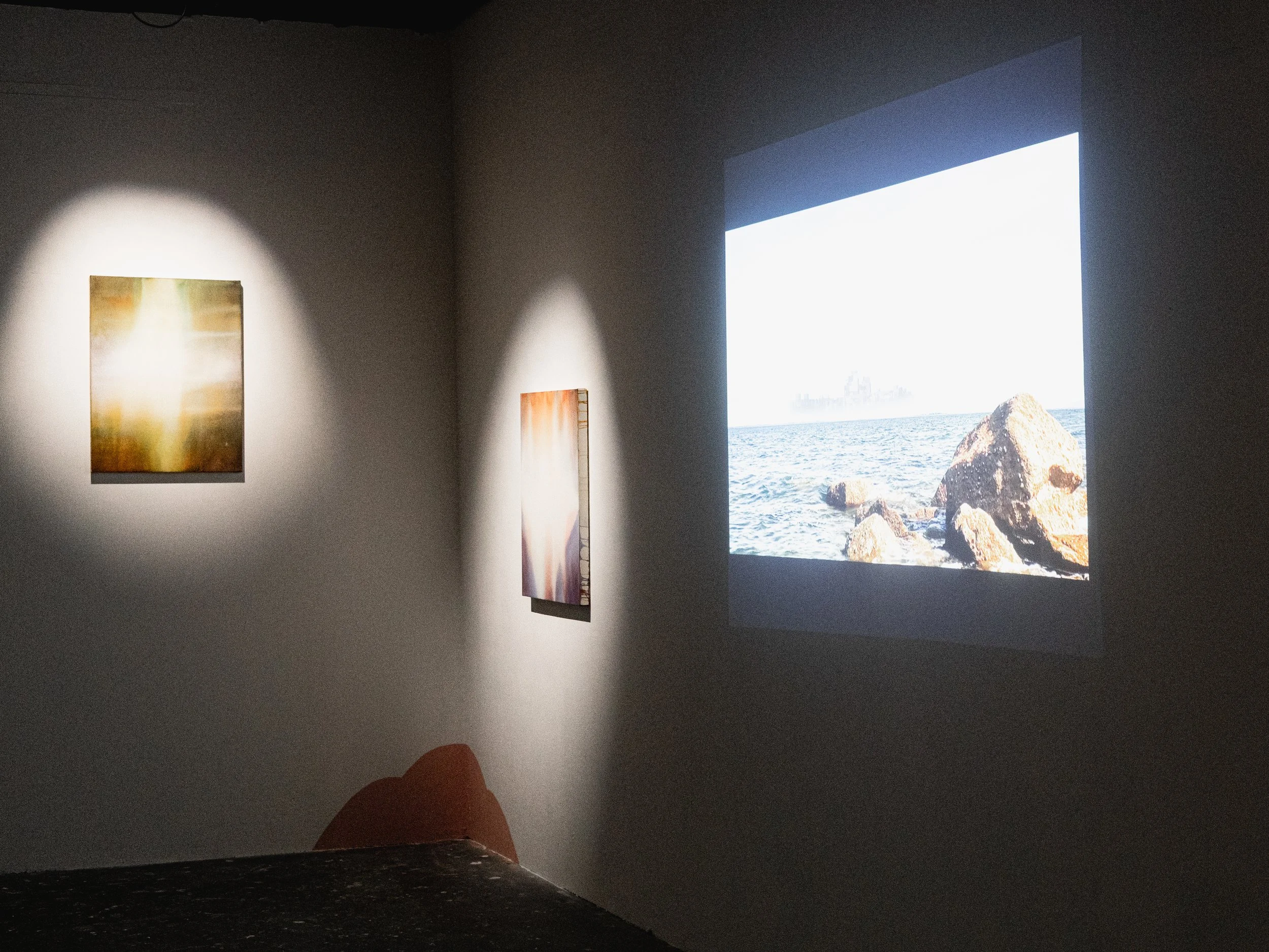 An art gallery with two paintings illuminated on a gray wall and a projection of a rocky seashore scene on the adjacent wall.