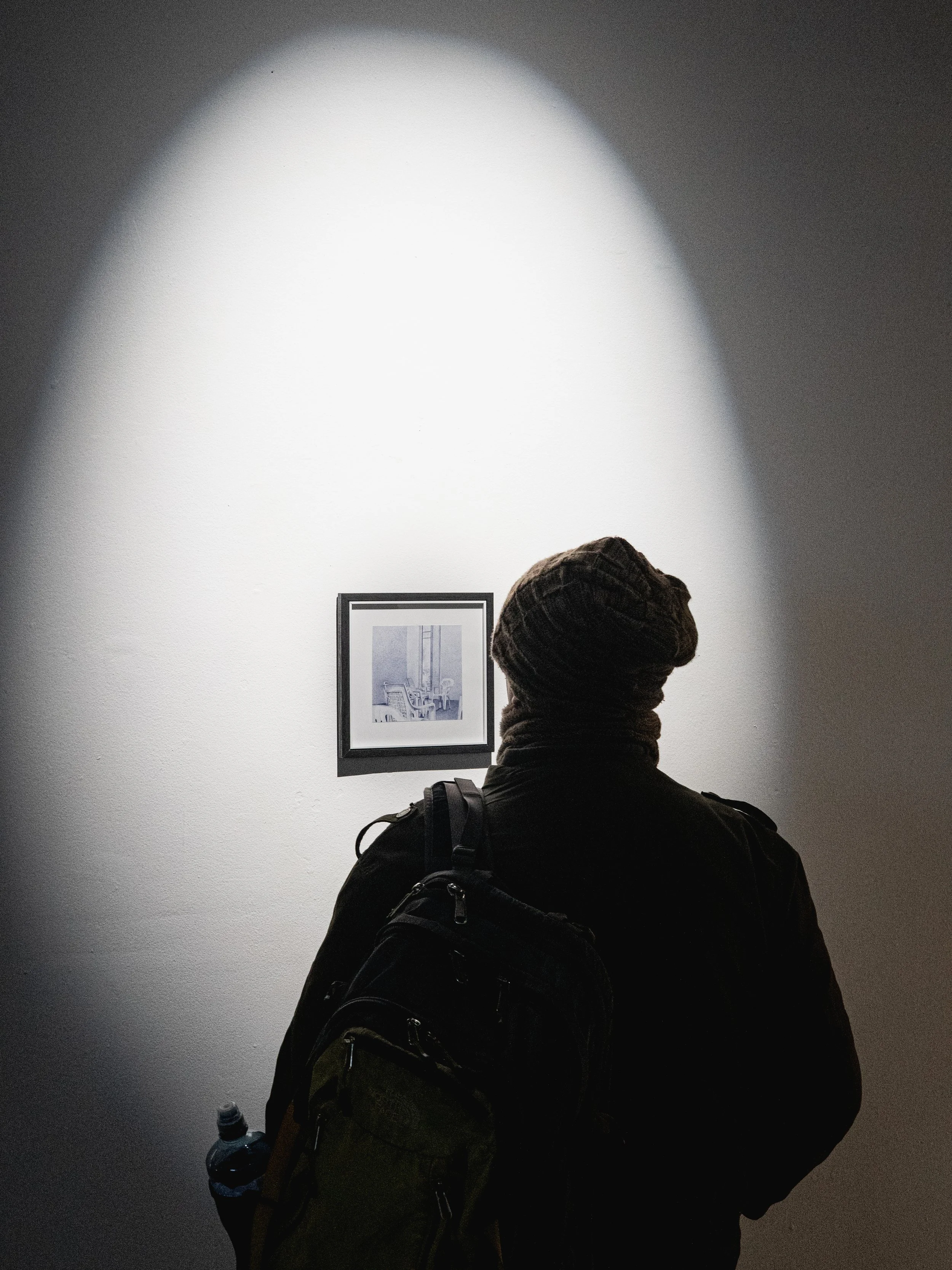 Person wearing a beanie and backpack looking at framed artwork on a wall in a dimly lit gallery or museum.
