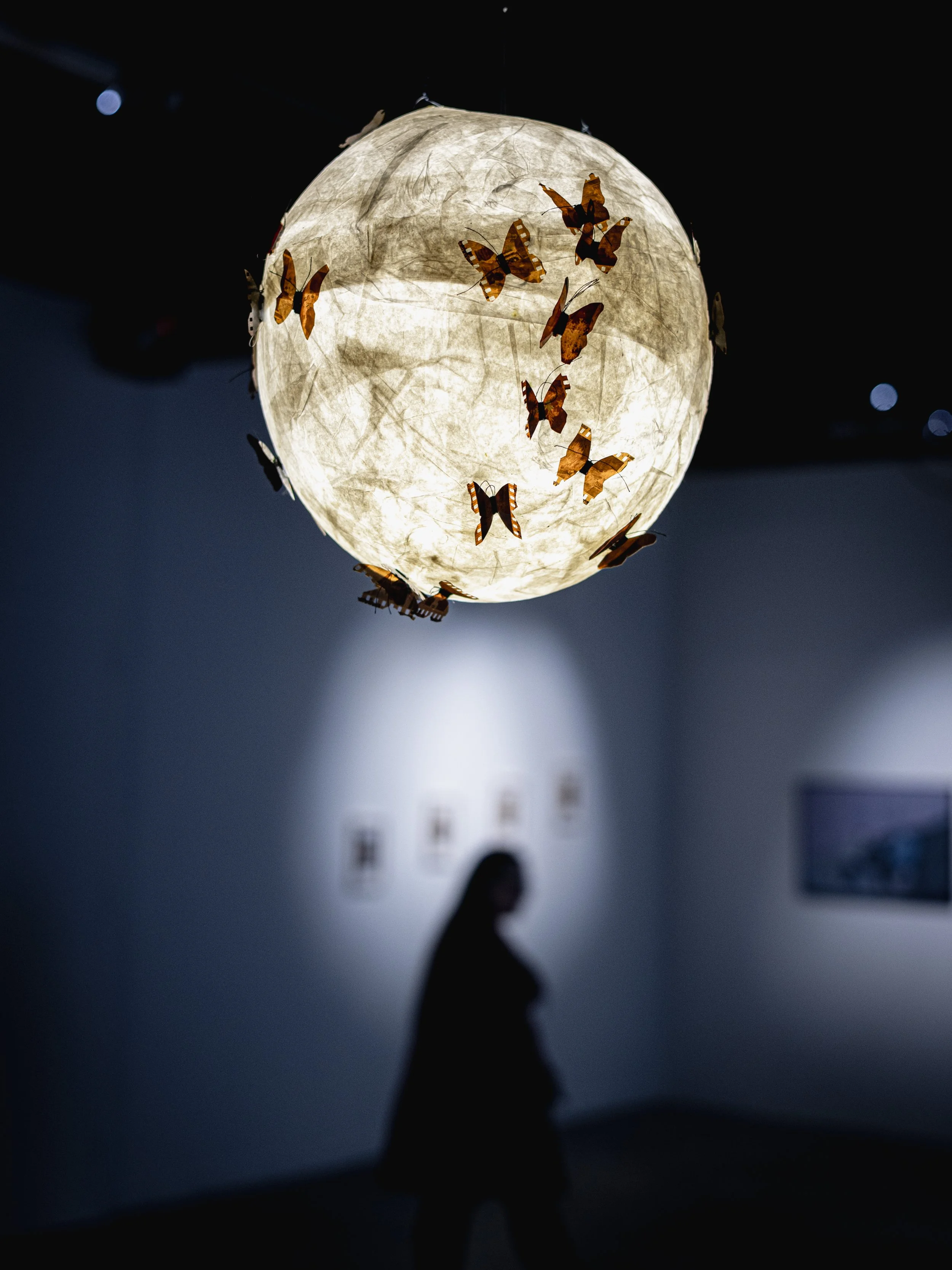 A woman in silhouette observes an art installation featuring a glowing paper lantern with brown and orange butterfly decorations, suspended from the ceiling in a dimly lit gallery.