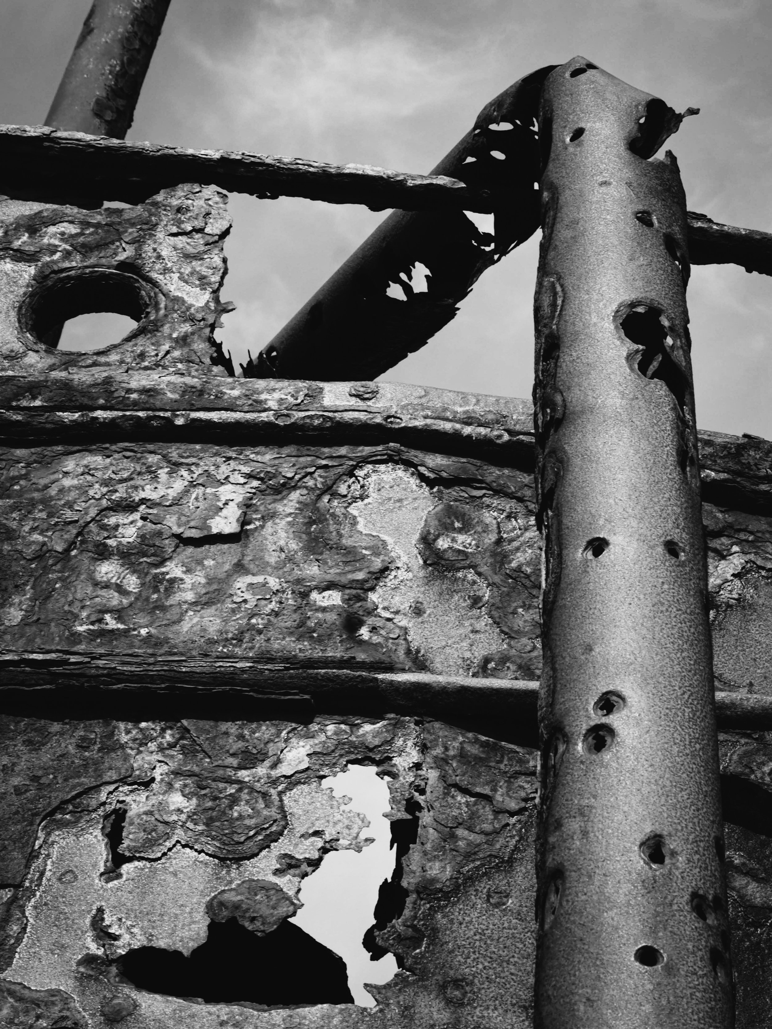 Close-up of rusted, deteriorated metal parts with holes and peeling surfaces, including a vertical pipe with multiple holes and a textured, corroded panel with additional holes, in black and white.