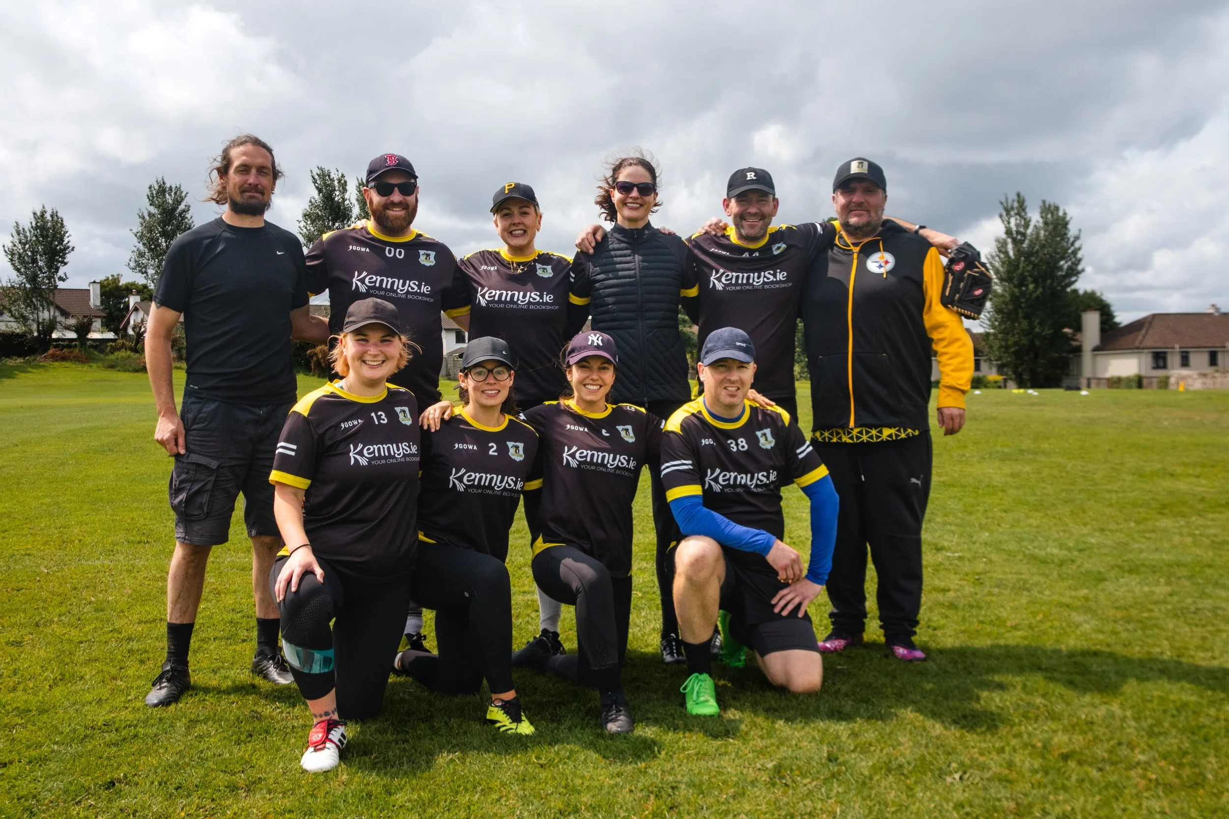 Group of people, including men and women, posing together on a grassy field, some wearing sports jerseys, under a cloudy sky.