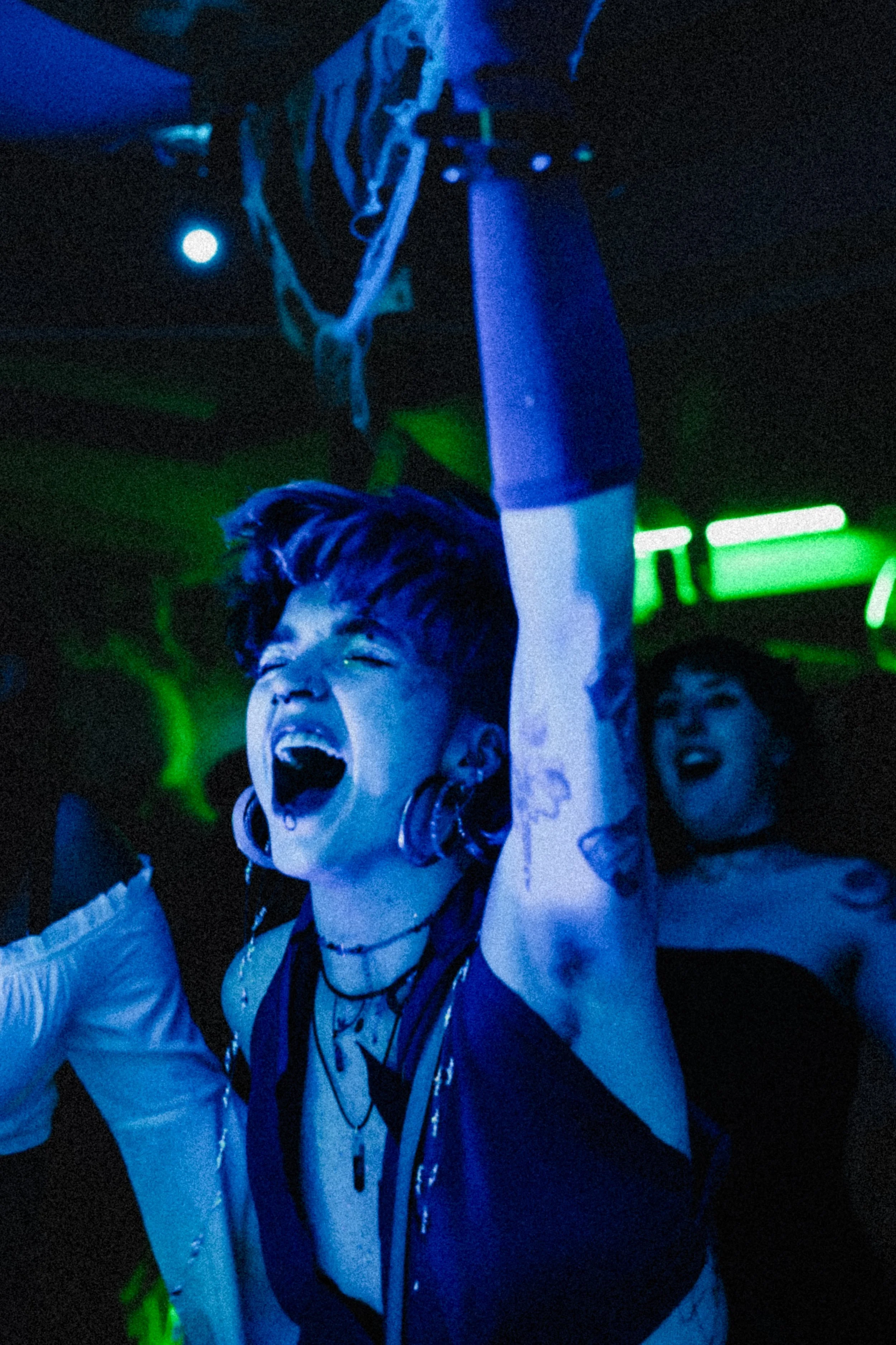 Woman with short dark hair, large earrings, and multiple necklaces dancing and singing passionately at a club, illuminated by blue and green neon lights, with other people in the background.