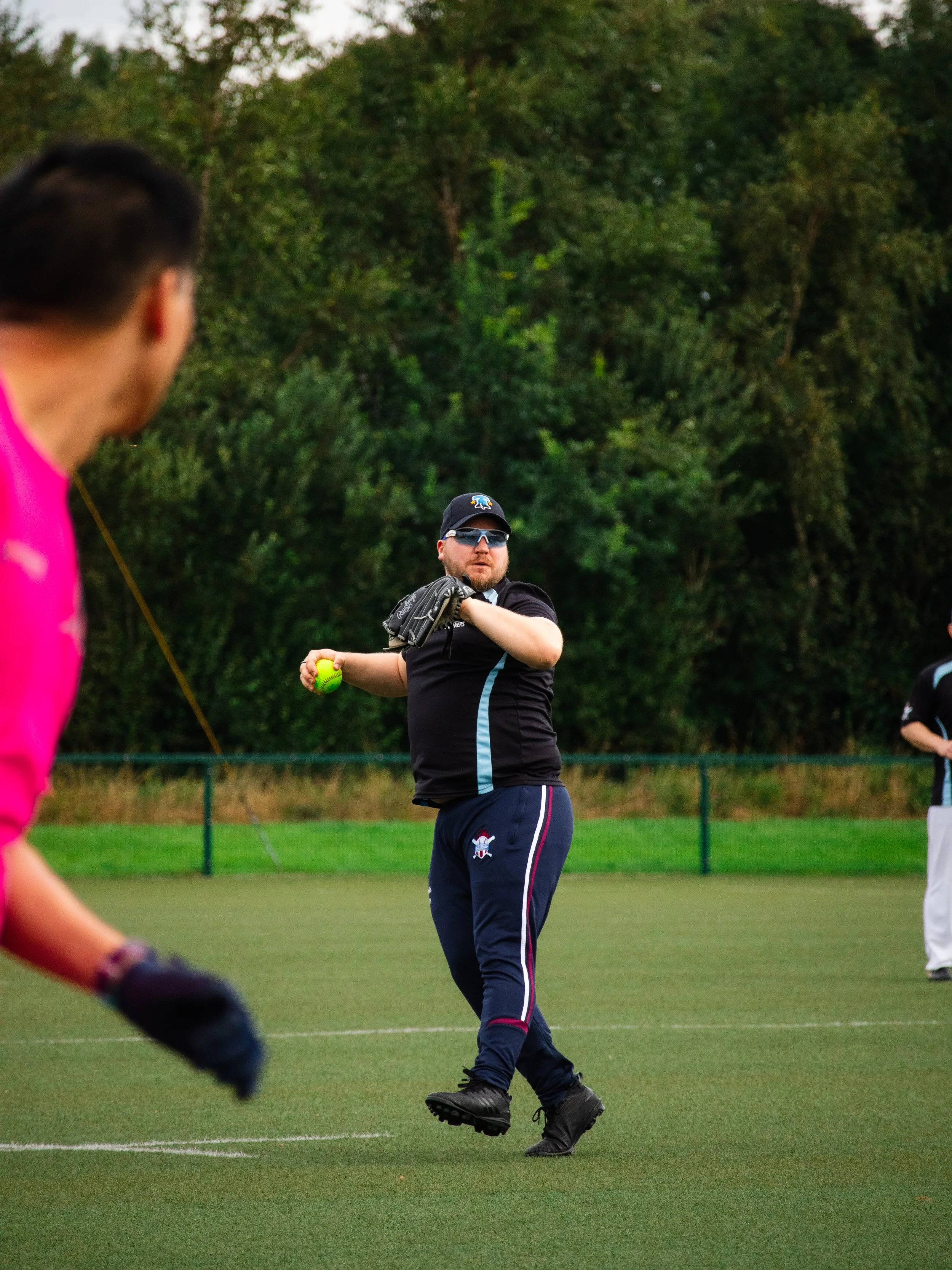 A man in a black shirt and navy athletic pants with red and white stripes is throwing a baseball on a grassy field. He is wearing sunglasses, a baseball cap, and a glove on his left hand. In the foreground, part of a person in a pink shirt and black 